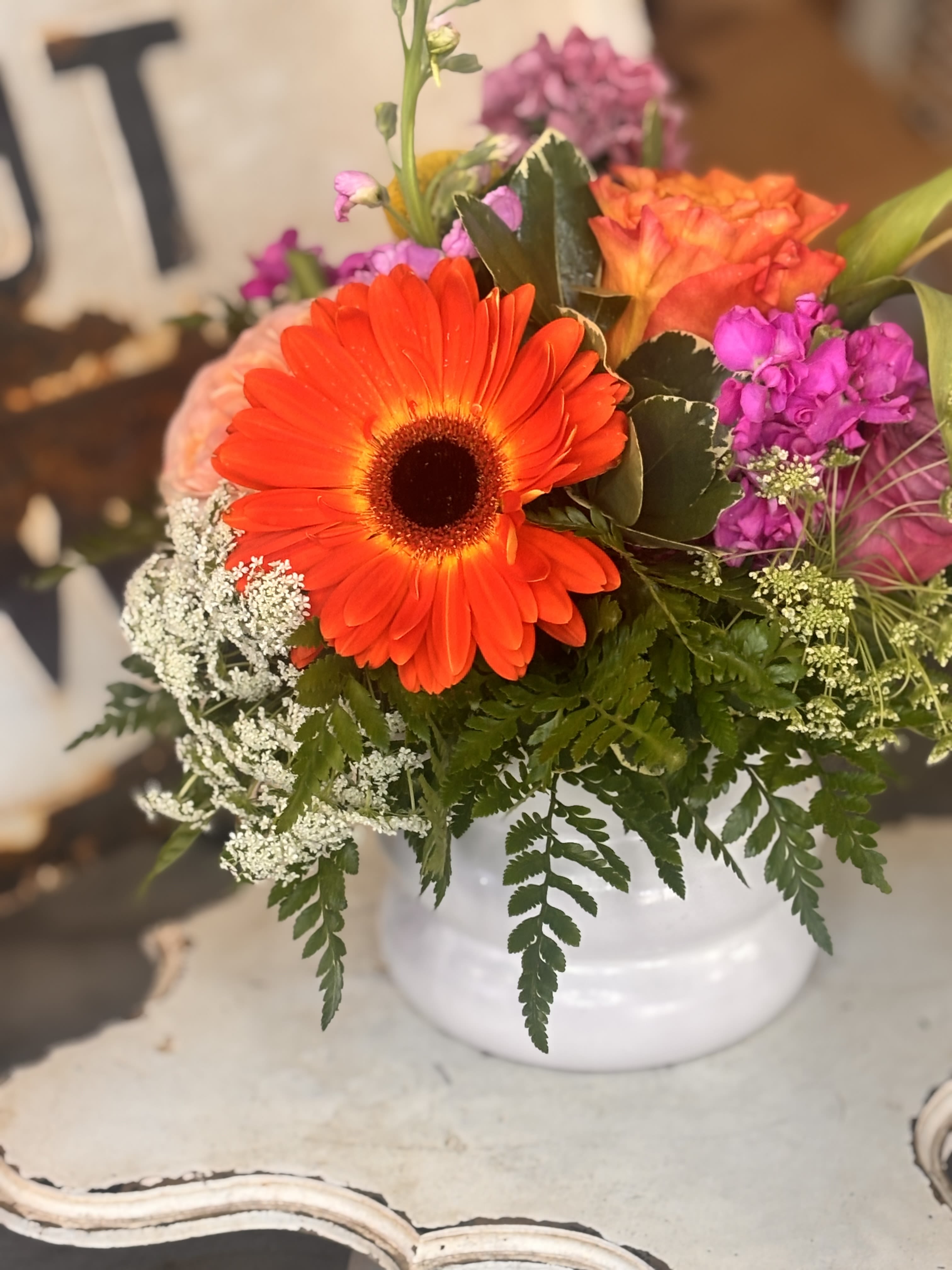 The Halle  - A vivid orange gerbera takes center stage in this cheerful mixed bouquet, making The Halle a perfect birthday bouquet, thank-you arrangement, or just-because gift for someone who loves bright, playful color. The lively mix of orange, pink, purple, white and yellow blooms reads joyful and modern - the kind of bouquet that makes the person you're thinking of feel noticed and celebrated the moment it arrives.  This garden-style arrangement pairs gerbera daisies, roses (including peach and lavender tones), tulips, carnations, clustered accent blossoms, and a dark rosette succulent for surprising texture, all softened by airy white filler flowers and fern-like greenery. The design is compact and rounded with layered petals and fleshy succulent leaves creating a pleasing contrast. It's presented in a clean white ceramic vase that gives a casual, polished finish and makes the bouquet easy to display.  Ideal for birthdays, congratulations, or brightening someone's day, this arrangement is made to feel fresh from the garden and ready to enjoy. Sizes are crafted to keep the composition full and balanced; for best longevity, refresh the water every two days and trim stems slightly when you change the water. Order with confidence for a colorful gift that reads upbeat and heartfelt.