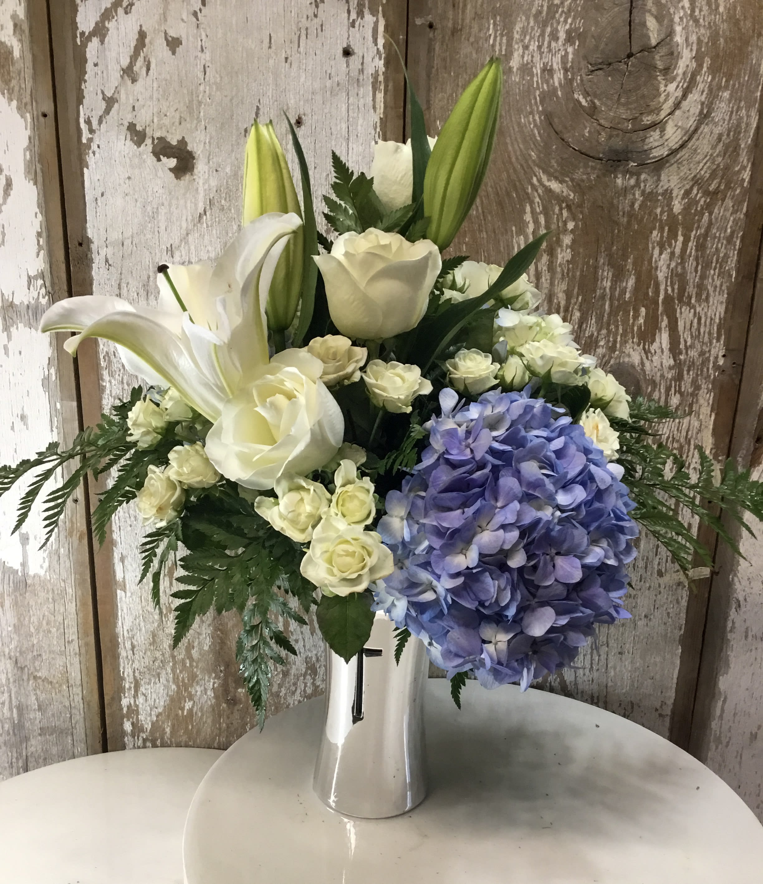 White lilies and roses with a blue hydrangea cluster in a silver vase.