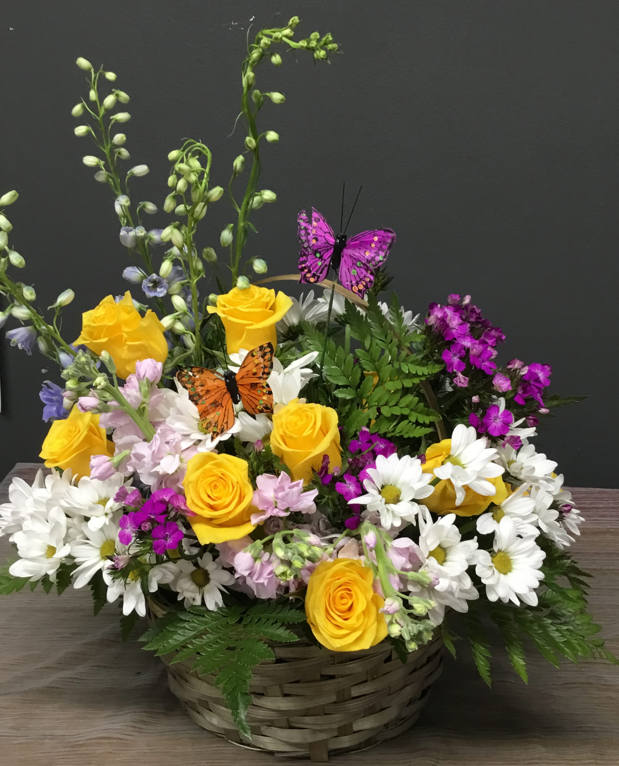Basket arrangement of yellow roses, white daisies, and purple flowers with butterfly picks