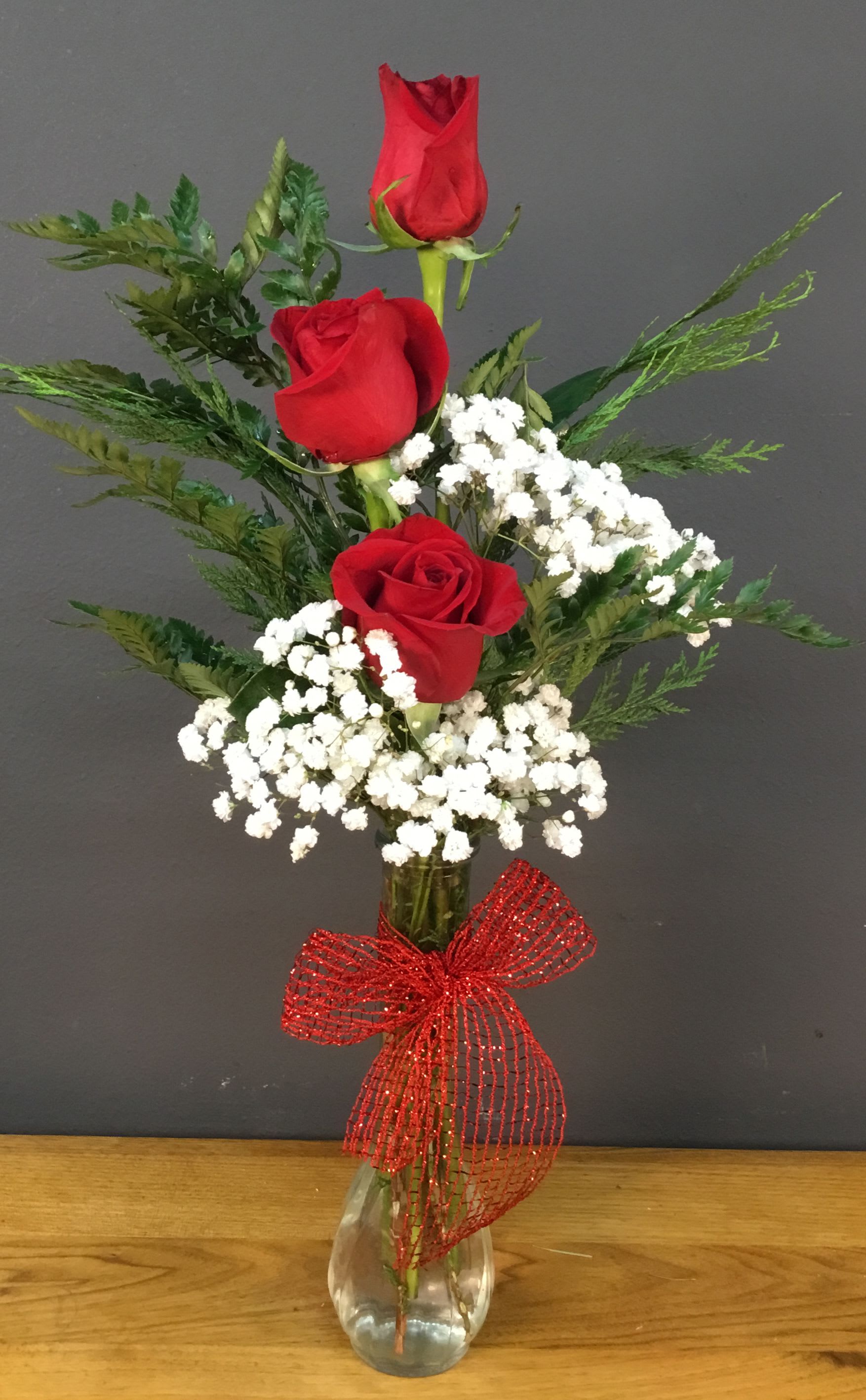 Three red roses in a clear vase with white baby's breath and a red bow