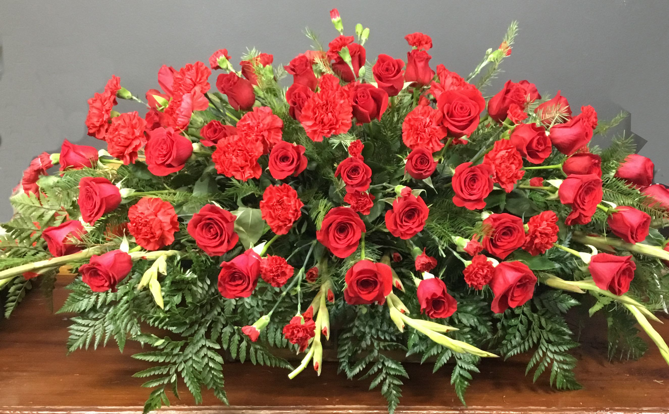 All Our Love Casket Spray - A dramatic, full-bodied casket spray that speaks of deep love and respect, the Love Casket Spray blends rich red roses and red carnations into a bold, formal tribute perfect for a funeral or a memorial honoring a beloved partner. This arrangement's intense red palette and low, broad profile create an immediate visual statement-ideal when you want the person you're remembering to feel cherished and honored.