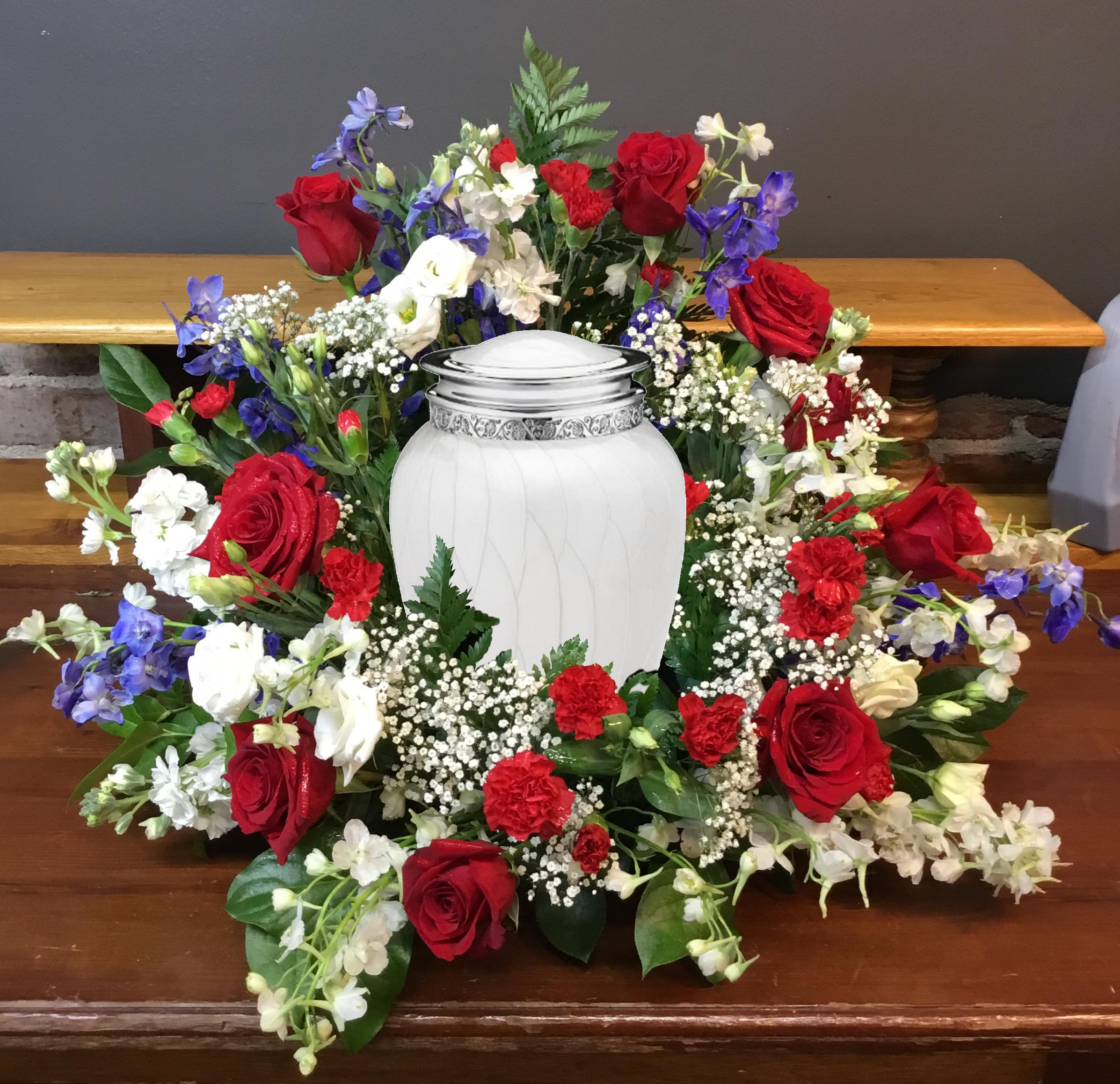 Patriotic Urn Piece - A formal circular sympathy arrangement presented around a white funeral urn, this patriotic urn piece combines red roses, white blooms and blue accents to create a dignified memorial tribute. Designed for a funeral or memorial service, the red-and-white palette reads ceremonial and respectful while touches of blue lend contrast and depth-making the person you're honoring feel remembered with quiet dignity.