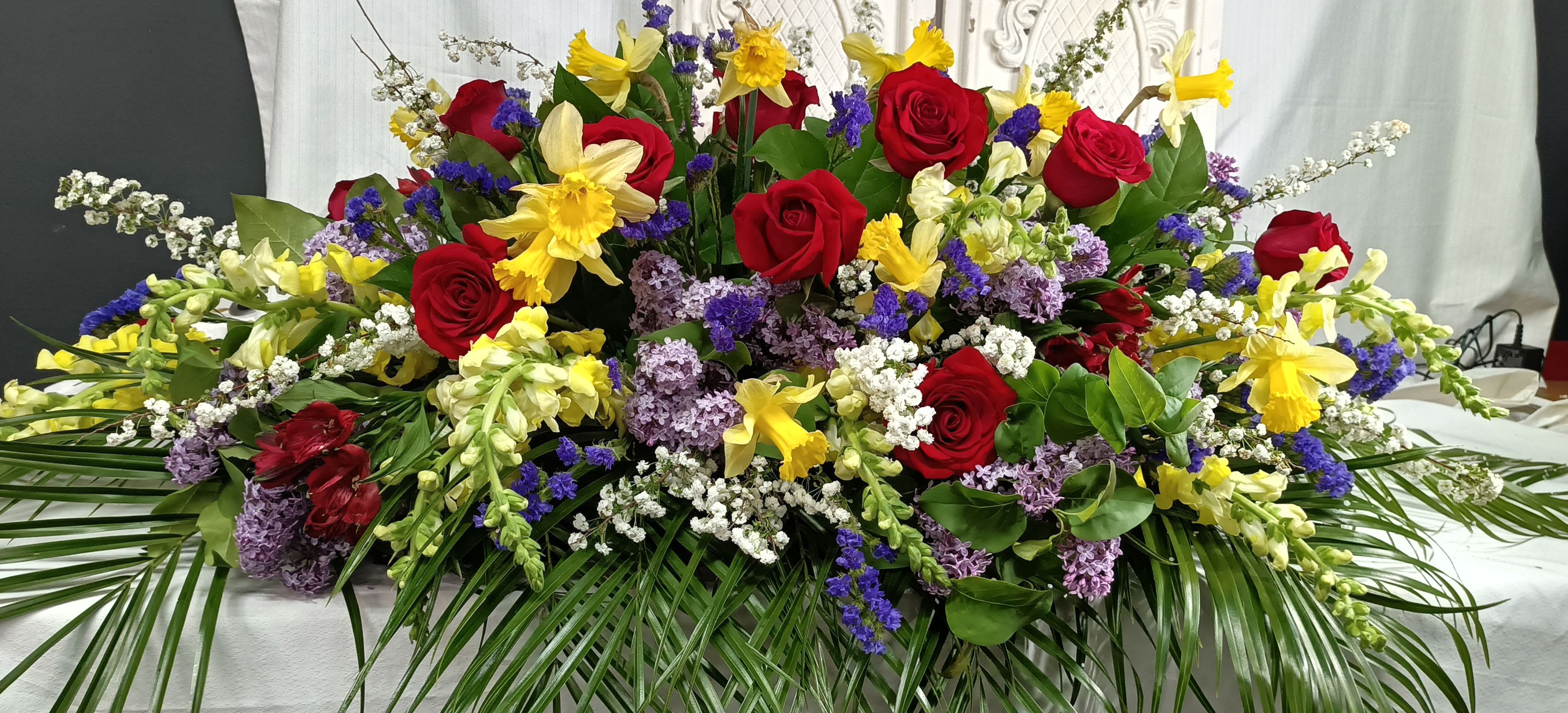 Jewel Tone Casket Spray - A bold, ceremonial floral spray that honors a life with jewel-toned drama-the perfect funeral or memorial casket spray to convey deep affection and respect. Clusters of red roses and bright yellow daffodils take center stage, while purple and white filler flowers add rich contrast and a sense of reverent beauty, creating a striking tribute for the person you're remembering.