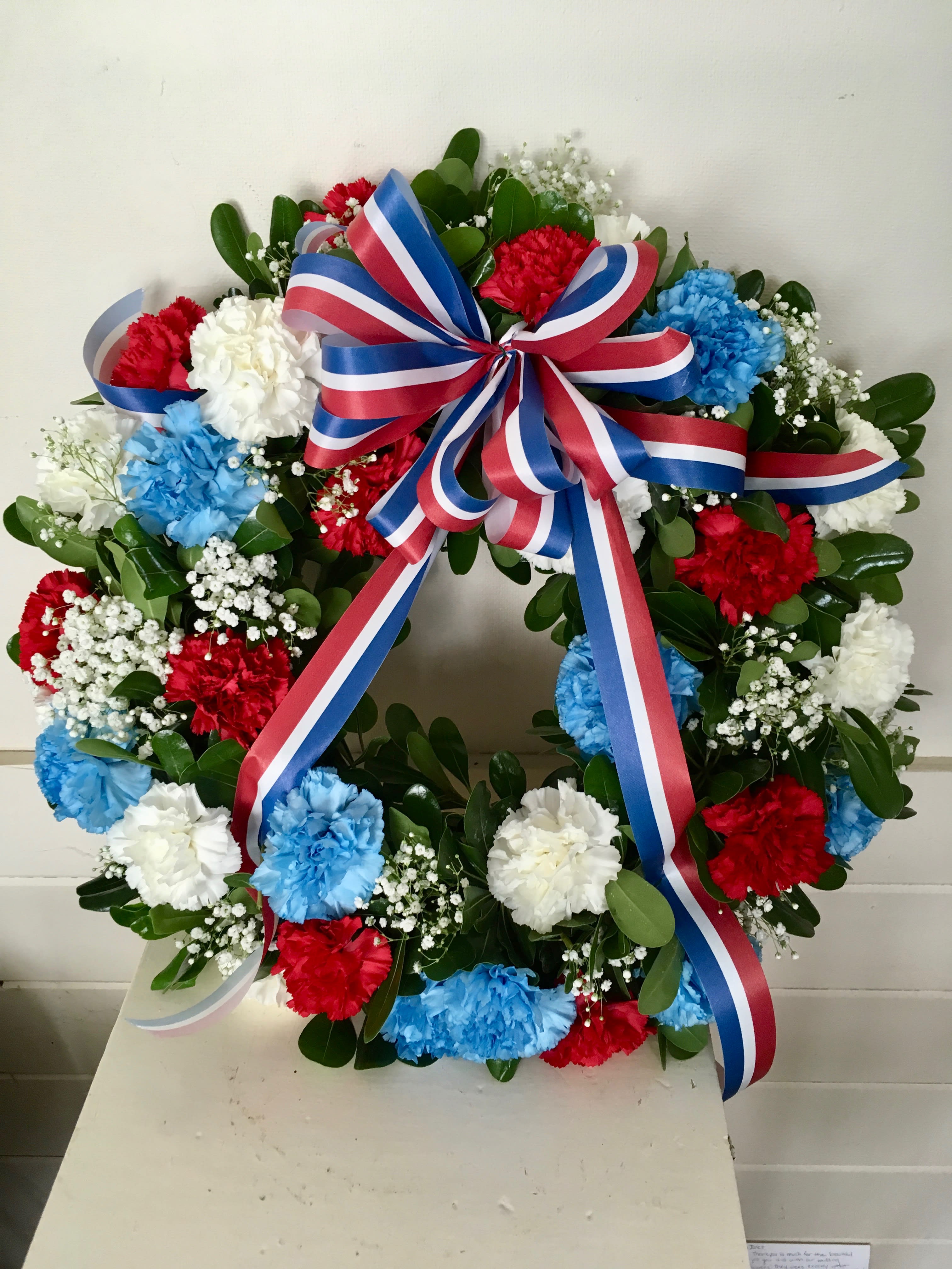 Round floral wreath with red, white, and blue carnations and a striped ribbon bow