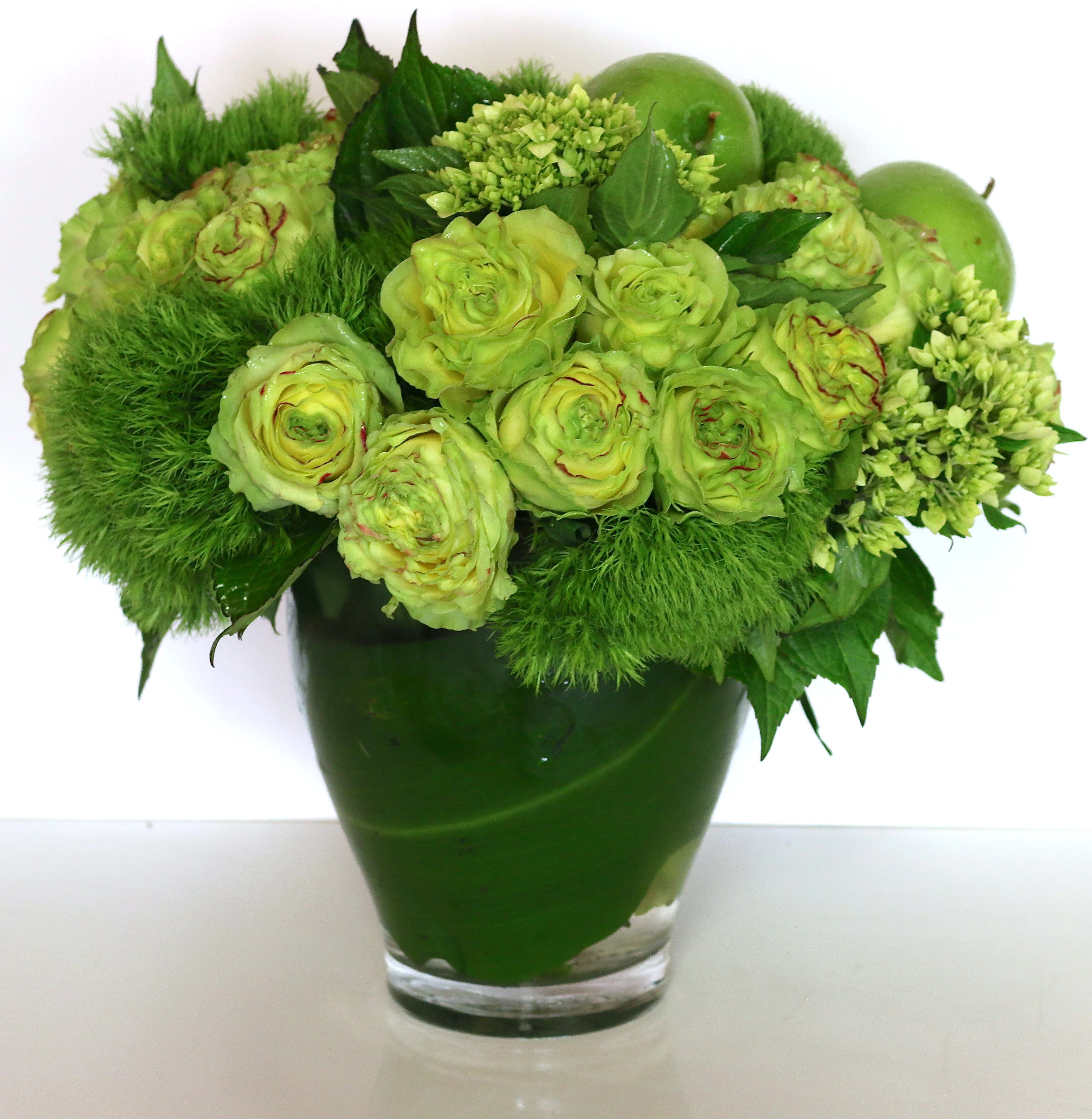 Green floral arrangement in a glass vase with round blooms and apples