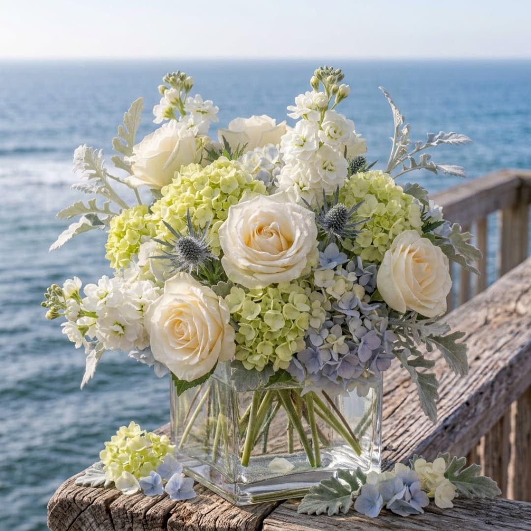 White roses and pale hydrangeas in a clear glass vase by the sea