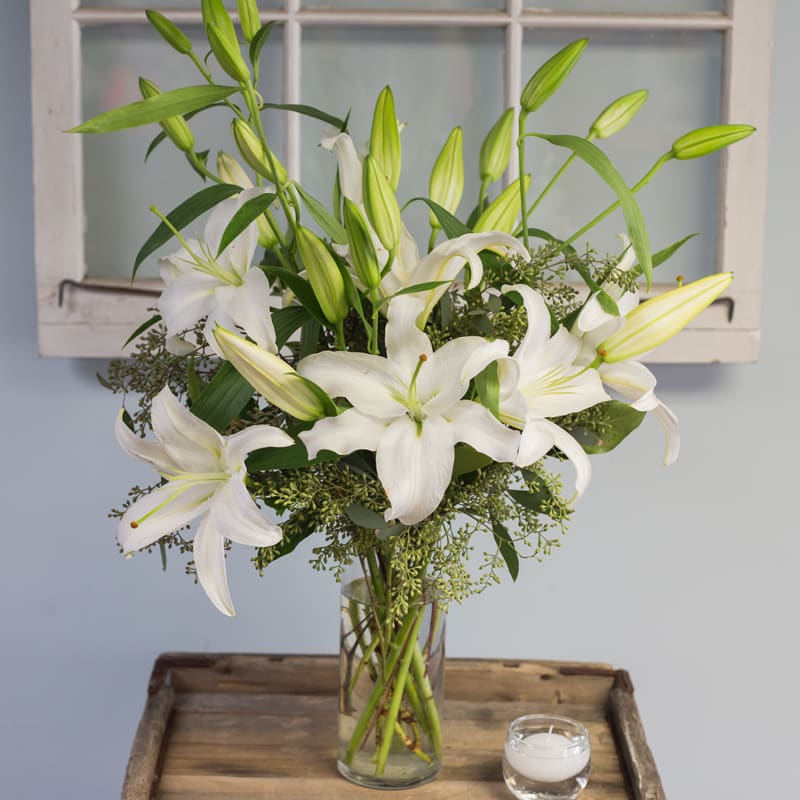 White lilies arranged in a clear glass vase with a small candle nearby.