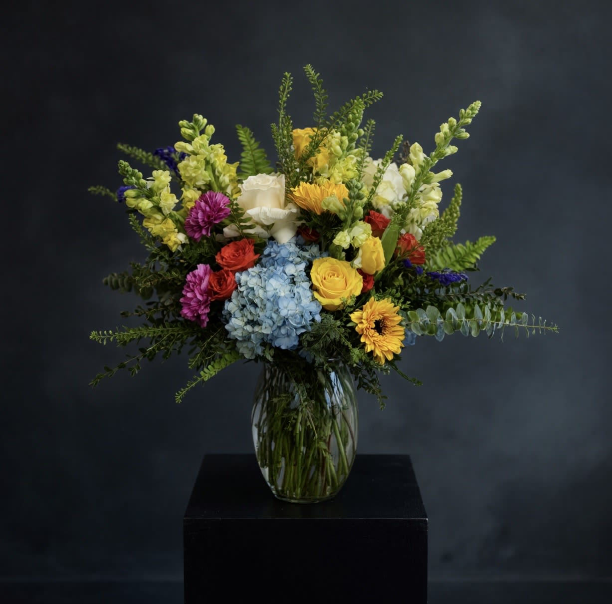 Sunshine And Grace - Bring joy and elegance into any space with the Sunshine &amp; Grace Bouquet—a radiant blend of hydrangeas, roses, sunflowers, tulips, snapdragons, ranunculus, stock, and lush greenery beautifully arranged in a glass vase. This stunning bouquet captures the essence of warmth, grace, and natural beauty, making it the perfect gift for any occasion.