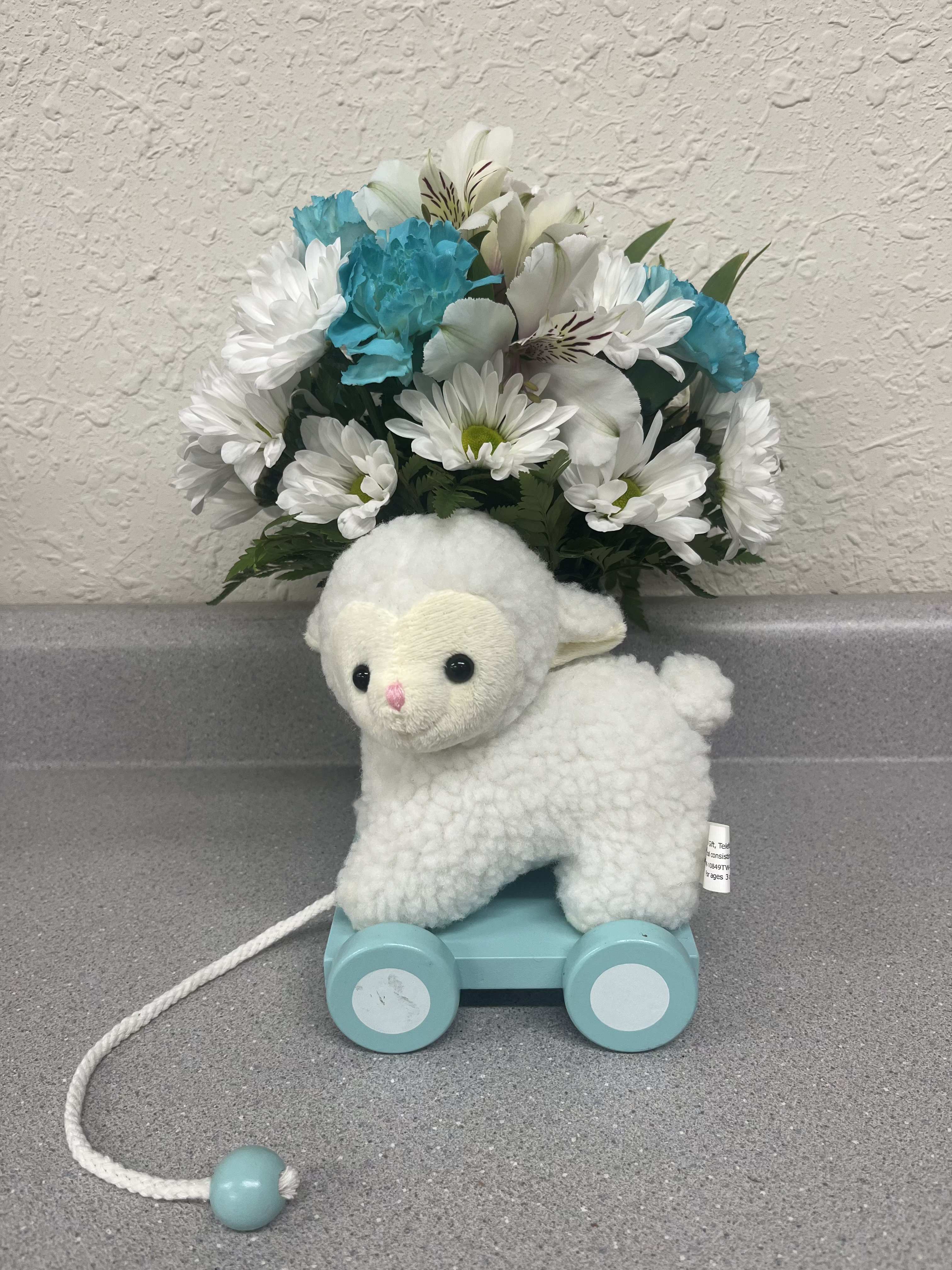 Plush lamb toy on a blue pull cart with white and blue flowers behind it