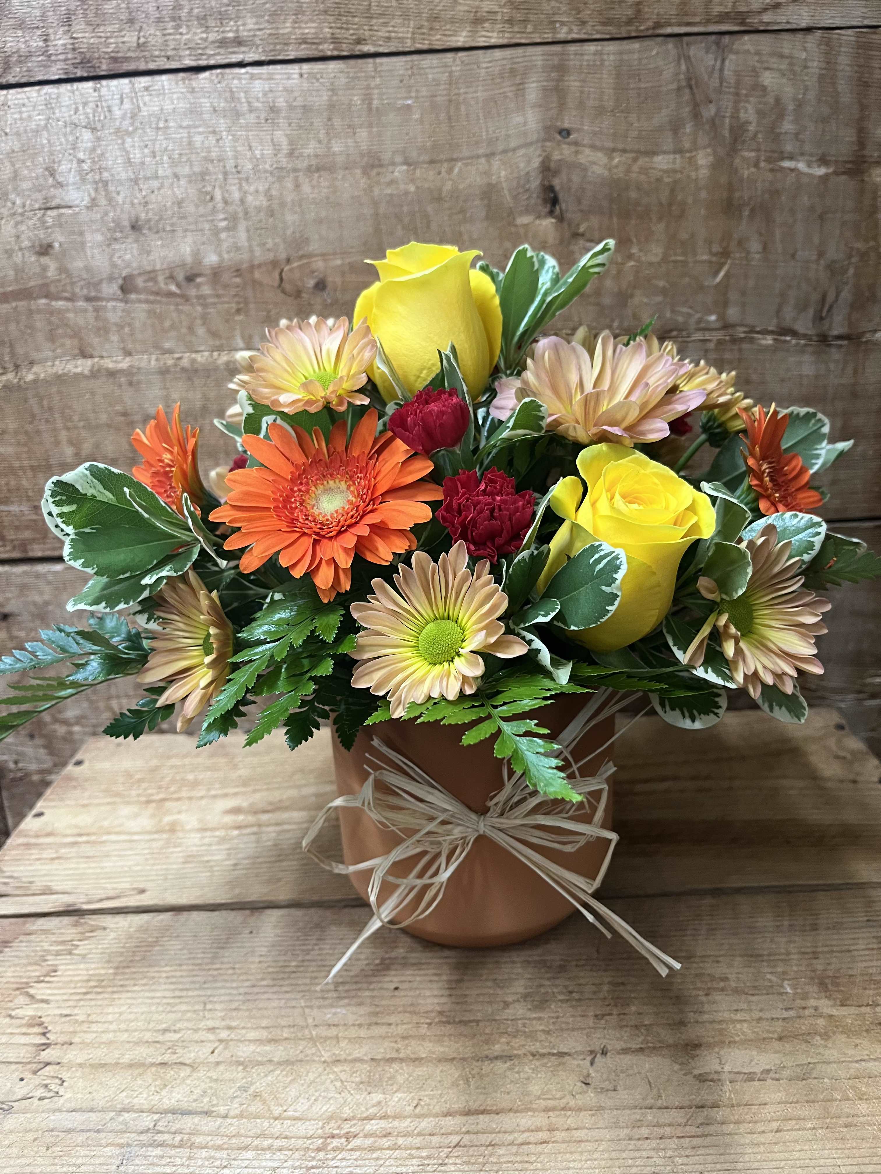 Terracotta Sunset - Bright, warm colors spill from a rustic terracotta pot in this cheerful flower arrangement. The mix of yellow roses, orange gerbera daisies, tequila sunrise daisies, and deep red miniature carnations reads sunny and lively, sending a message of warmth and celebration to the person you're thinking of. It is perfect for Mother's Day, a birthday bouquet, a thank-you gift, or a just-because surprise.  Flowers sit densely in a compact, rounded design with variegated foliage for texture, finished with a raffia bow for a hand-tied, garden-style look. The arrangement is sized for tabletop display and has adorable rustic charm.