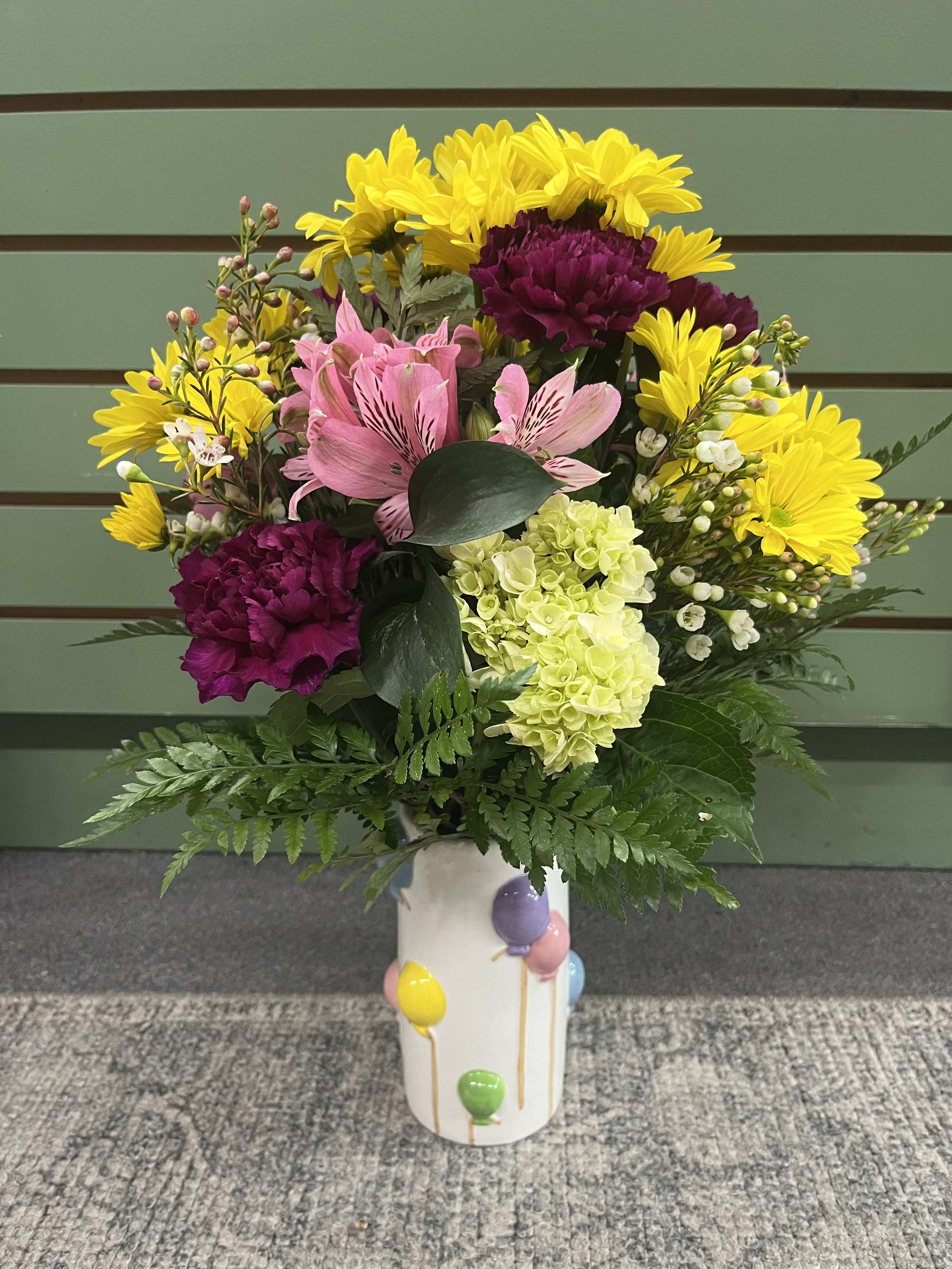 Celebration Time  - Bright, confetti-like color greets them the moment this bouquet arrives. As a birthday arrangement or a "just because" it reads joyful and celebratory. The white ceramic vase printed with tiny balloon motifs underscores the festive message for birthday or a congratulatory surprise. There are cheerful yellow daisies, soft pink alstroemeria, rich purple carnations, and a pale green hydrangea.   -Happy Birthday pick can be added if requested!   Perfect for sending birthday wishes, saying congrats, or brightening a friend's day in spring and beyond. They'll feel genuinely celebrated when these arrive. Standard care: keep in fresh water, trim stems every few days, and display away from direct heat to prolong bloom life.