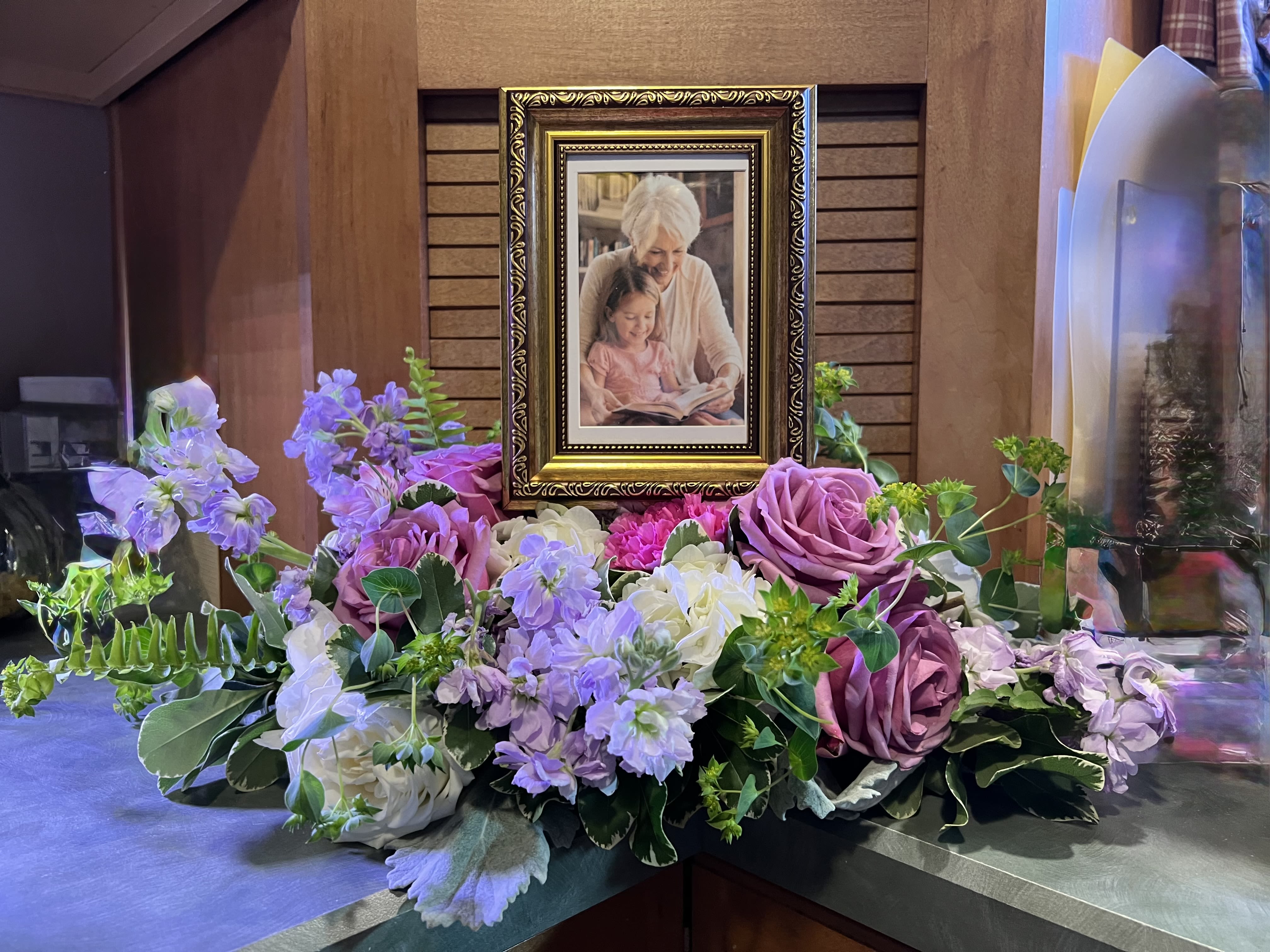 Surrounded with Flowers  - Soft lavender roses and whisper-pink blooms form a low, spreading tribute that gently surrounds either one or two framed photographs - an understated sympathy arrangement designed for memorial tables and remembrance services. The palette of purple, white, pink, and green reads tender and reflective; the composition places the portrait as the focal point so the person you're honoring feels seen and celebrated.  Large rose blooms anchor the design while carnations, stock, hydrangea-like clusters, daisies, and dianthus provide layered texture and airy filler across a broad horizontal base. Mixed greenery and small purple filler flowers soften the edges and add a garden-fresh, lush look. Sized to sit on a draped table, this formal yet personal piece integrates frames into the flowers for a cohesive presentation; it's intended for sympathy and memorial use and arrives ready to place beside photographs for an immediate, comforting display.