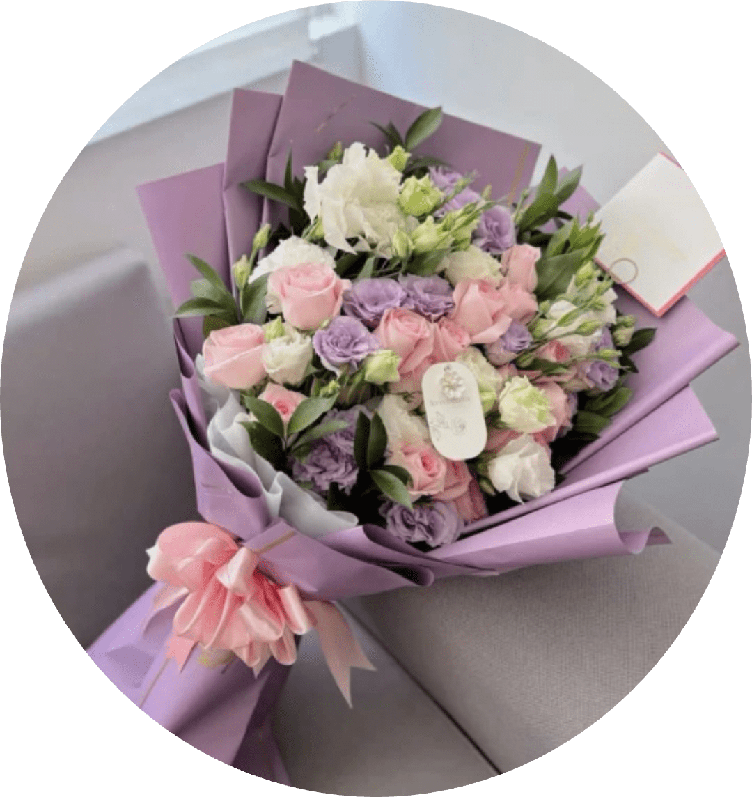 DOLORES - Lavender-toned blooms mingle with soft pink and creamy white roses in a rounded, full arrangement that reads like a pastel love letter - perfect as a birthday bouquet, anniversary flowers, or a just-because gift to make someone feel truly seen. The gentle mix of roses and lisianthus creates a romantic, feminine impression that will brighten a spring table or soften an everyday moment for the person you're celebrating.  This arrangement is wrapped in layered purple decorative paper with a pale pink satin ribbon and finished with a gift tag and greeting card for a polished, ready-to-give presentation. Small clusters of blossoms give the bouquet a textured, hand-gathered look; the color palette of purple, pink, white, and green keeps the feel fresh and delicate without being fussy.  Choose this as a thoughtful way to mark birthdays, anniversaries, or to send warm congratulations - she'll feel cherished the moment the bouquet arrives. Care is simple: keep stems in fresh water, trim ends before arranging in a vase, and place away from direct heat to extend vase life; comes wrapped and ready for delivery.