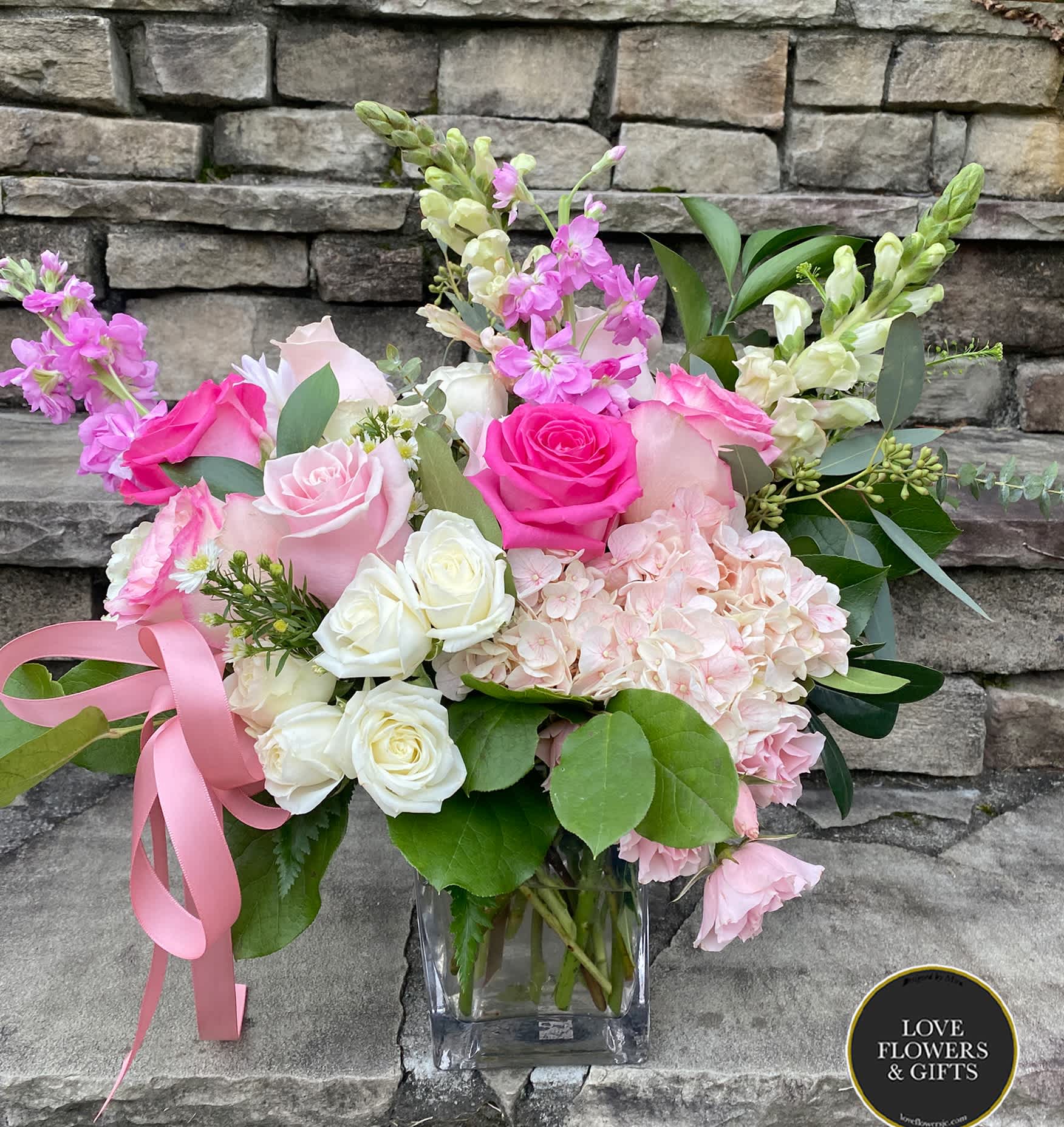 Pink and white roses, hydrangeas, and snapdragons in a clear glass cube vase with a pink ribbon.