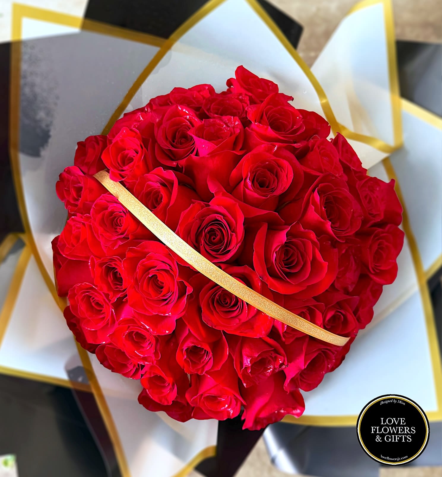 Round bouquet of tightly clustered red roses wrapped in white and black paper with gold accents.