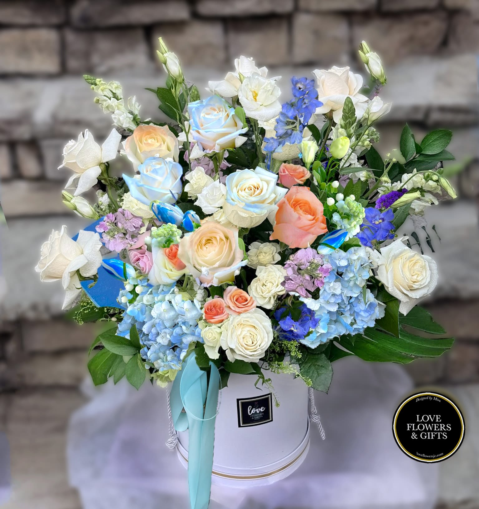 Pastel roses, hydrangeas, and mixed blooms arranged in a white hat box with aqua ribbon.