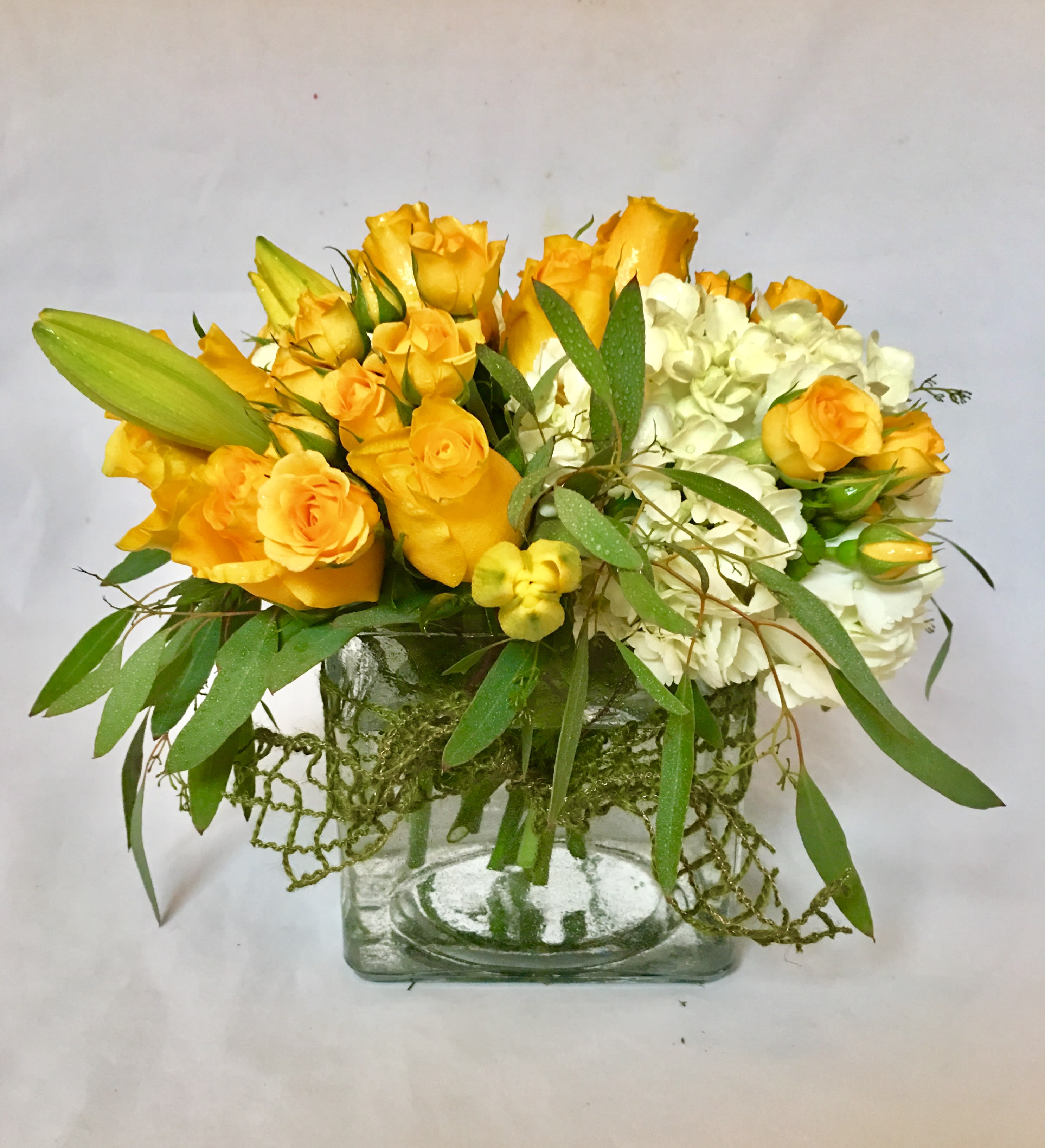 Low arrangement of yellow roses, lily buds, and white hydrangea in a clear glass cube vase