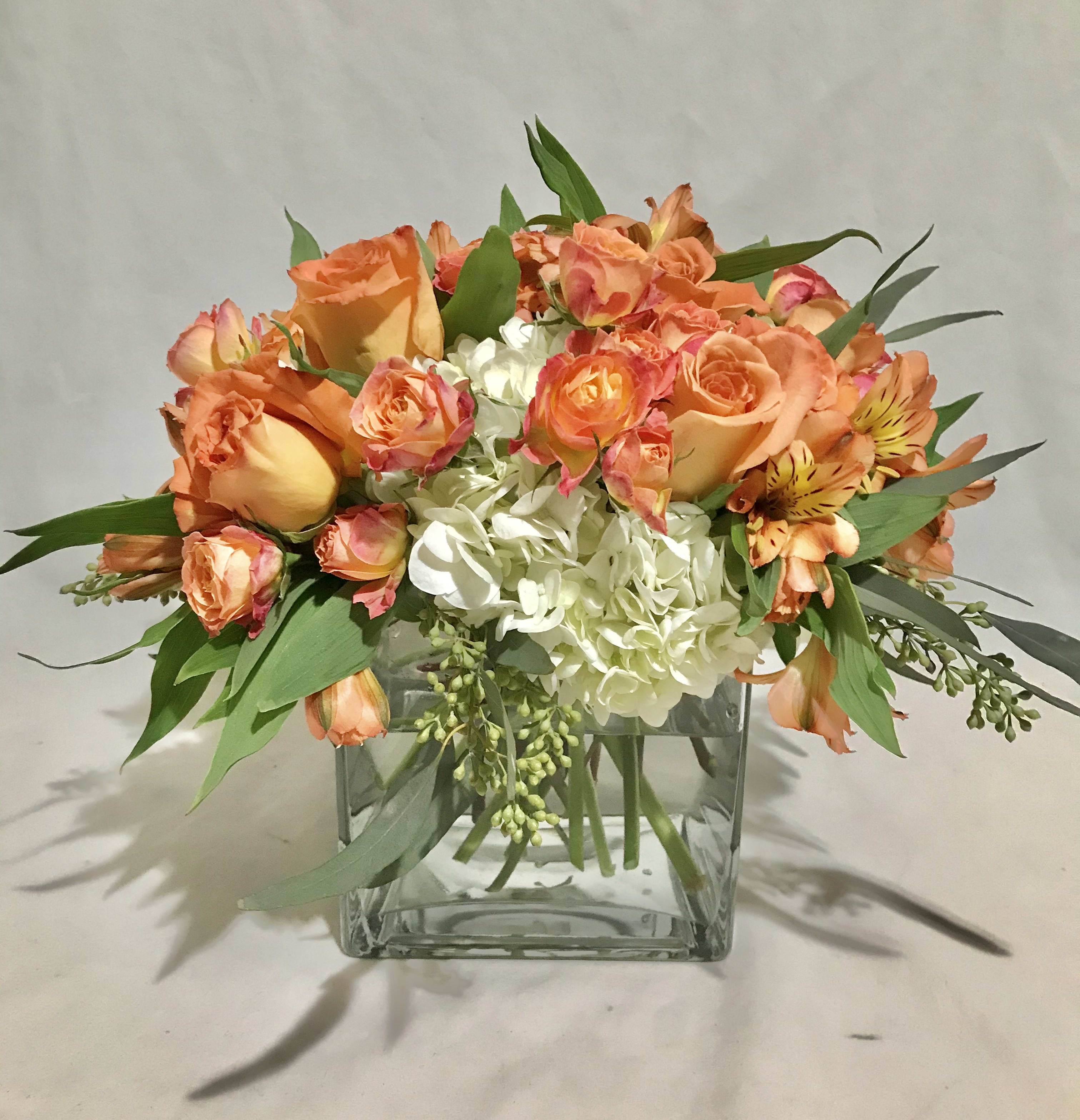 Short arrangement of orange roses and white hydrangeas in a clear glass cube vase