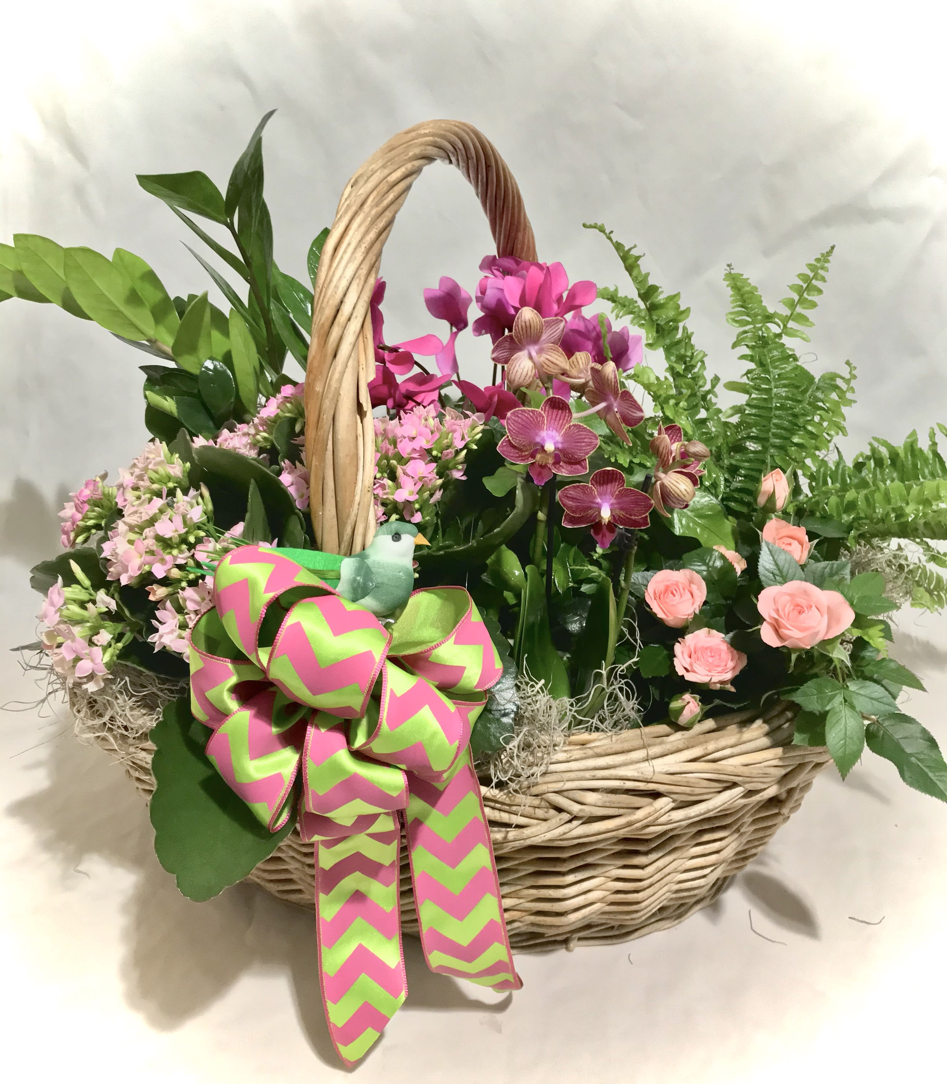 Basket of pink orchids, peach roses, and blooming plants with a large green and pink bow and small bird accent
