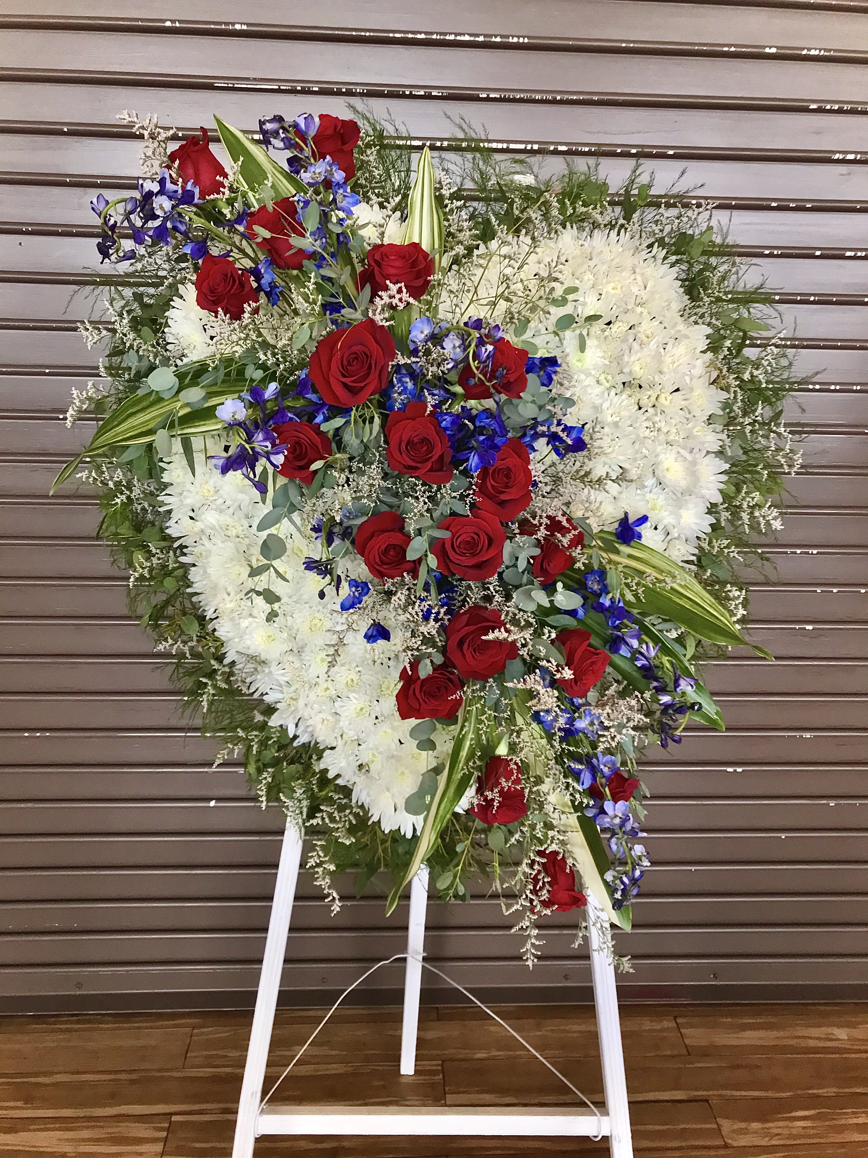 Tall heart-shaped standing spray with white flowers, red roses, and blue blooms on a white easel.