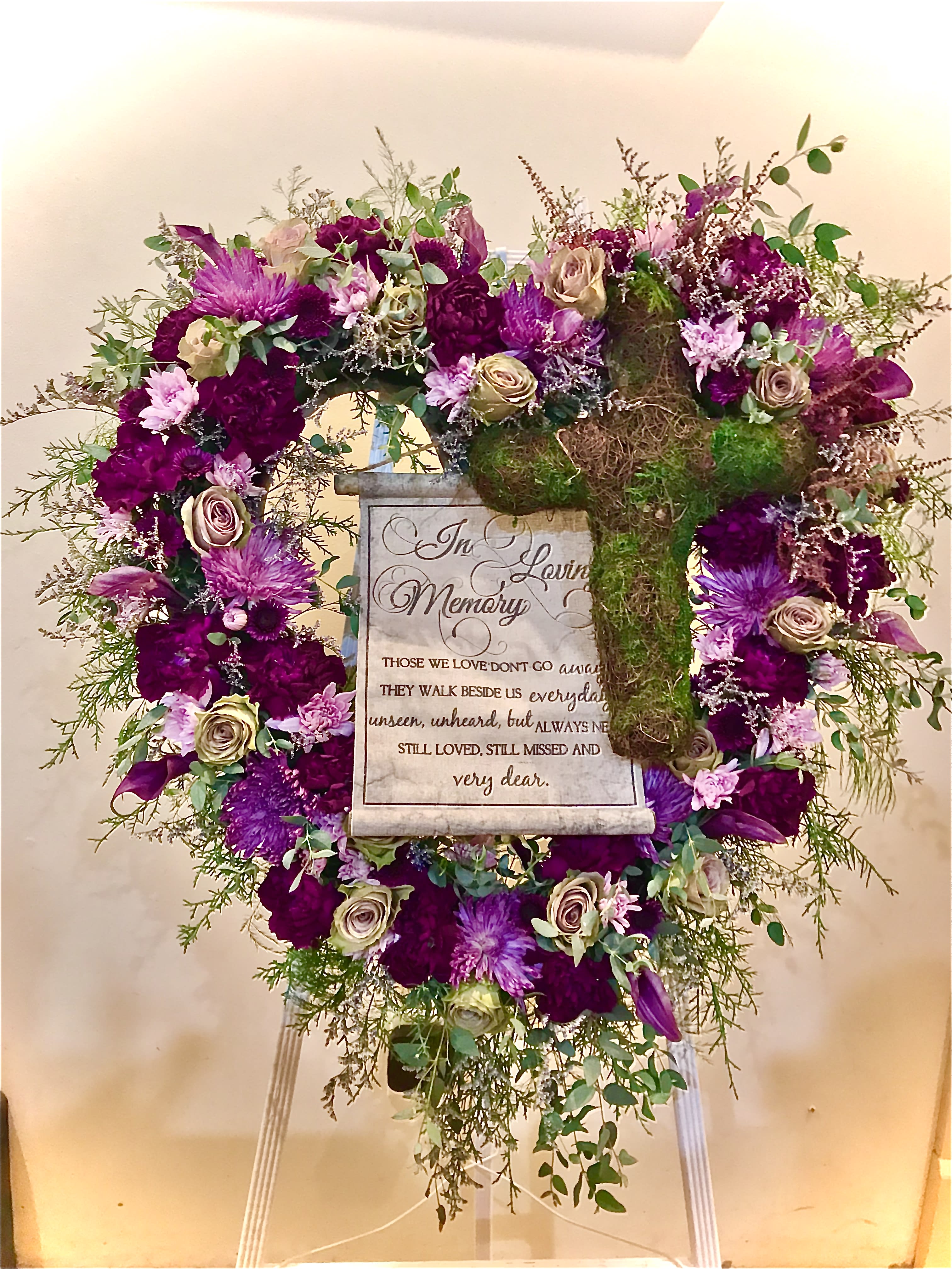 Standing sympathy wreath of purple and cream flowers with a moss-covered cross and memorial plaque on an easel