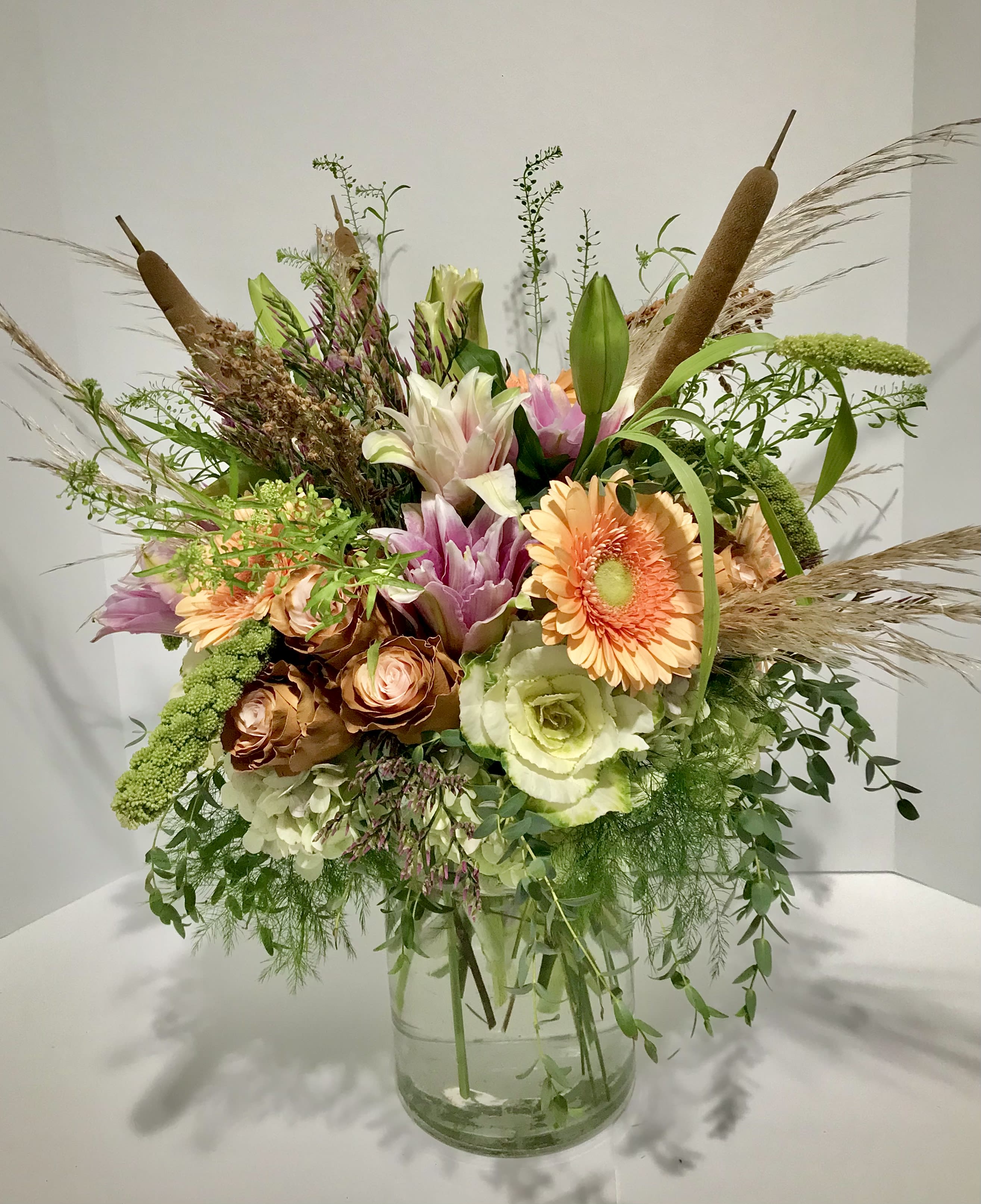 Tall rustic bouquet of peach gerbera, roses, lilies and white hydrangea in a glass vase with cattails and grasses.