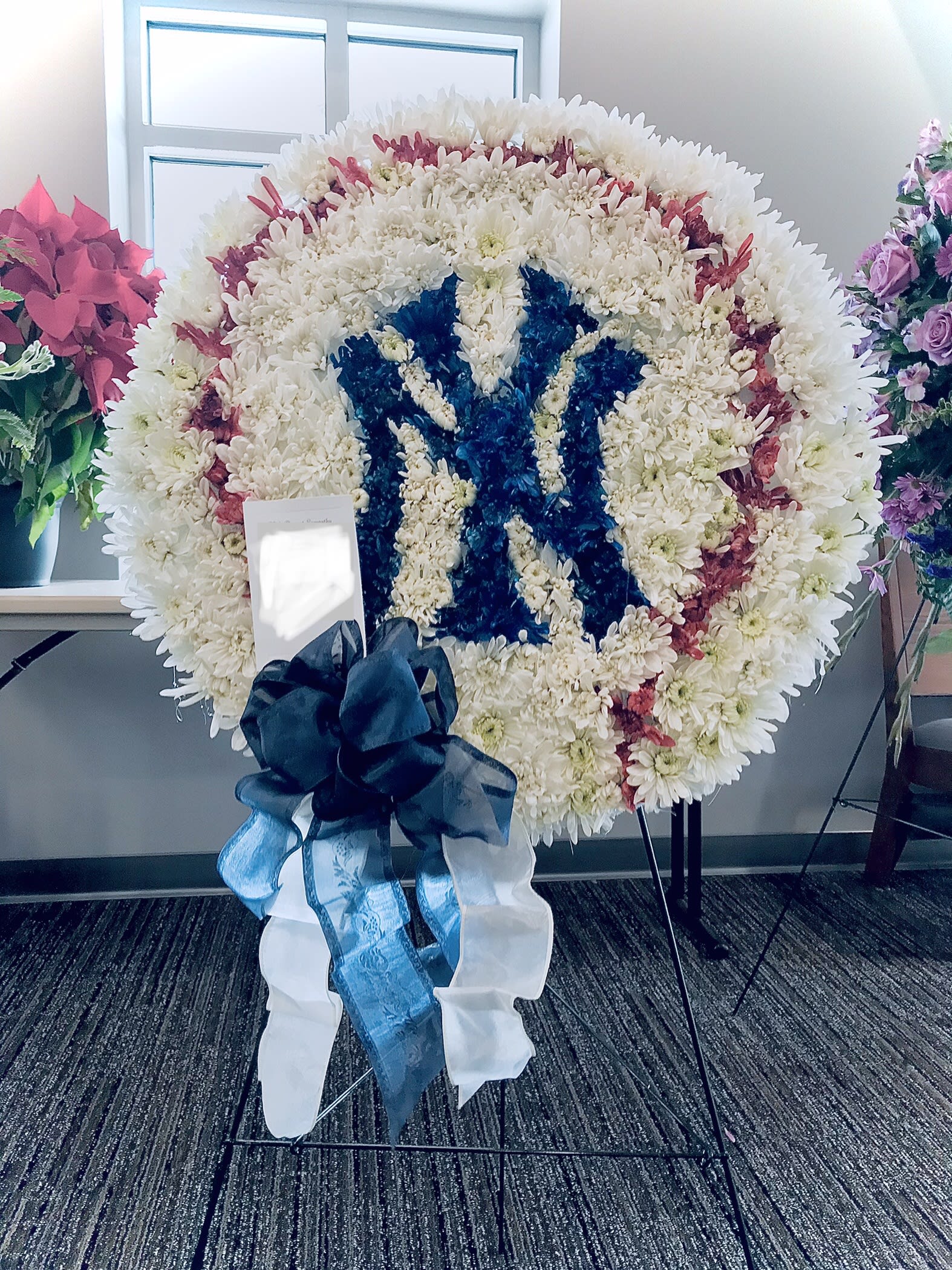 Large round standing wreath of white flowers with red accents and a blue floral emblem, finished with a dark bow.