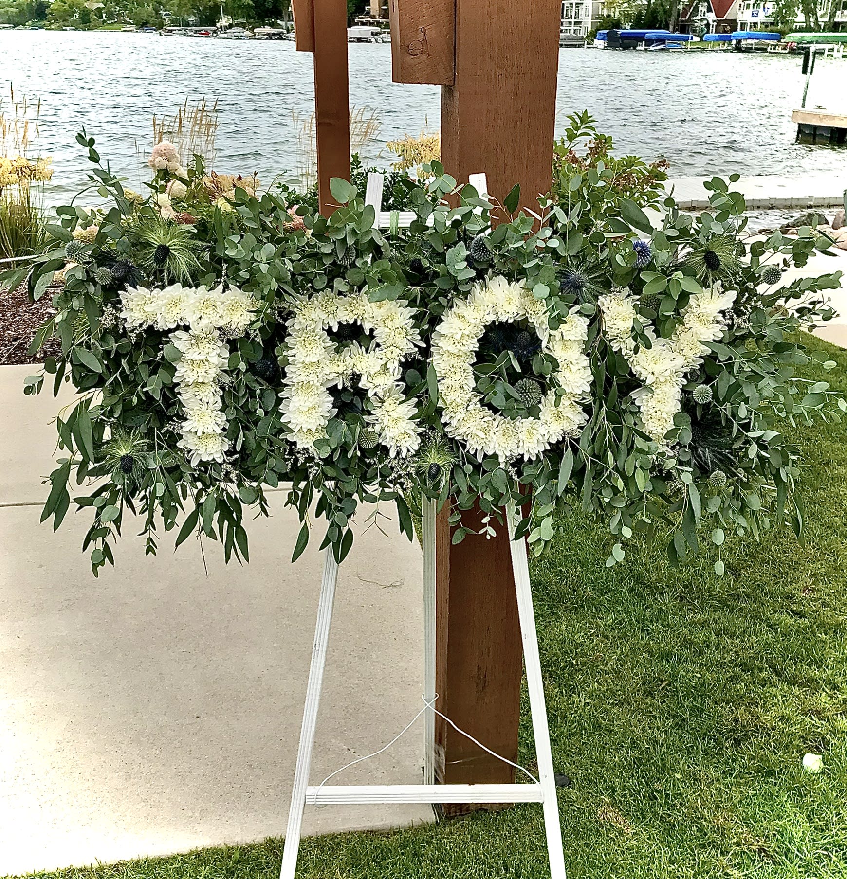 Standing tribute spray spelling TROY in white flowers with lush greenery on an easel