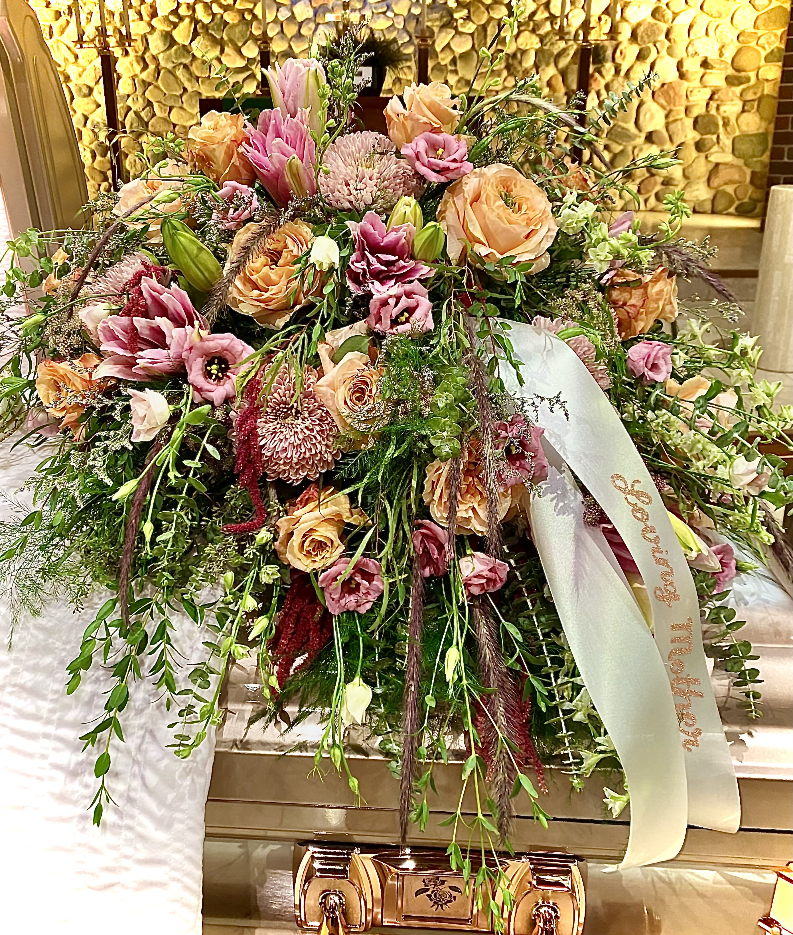 Large peach and pink casket spray with roses, lilies, and mums draping over a bronze casket