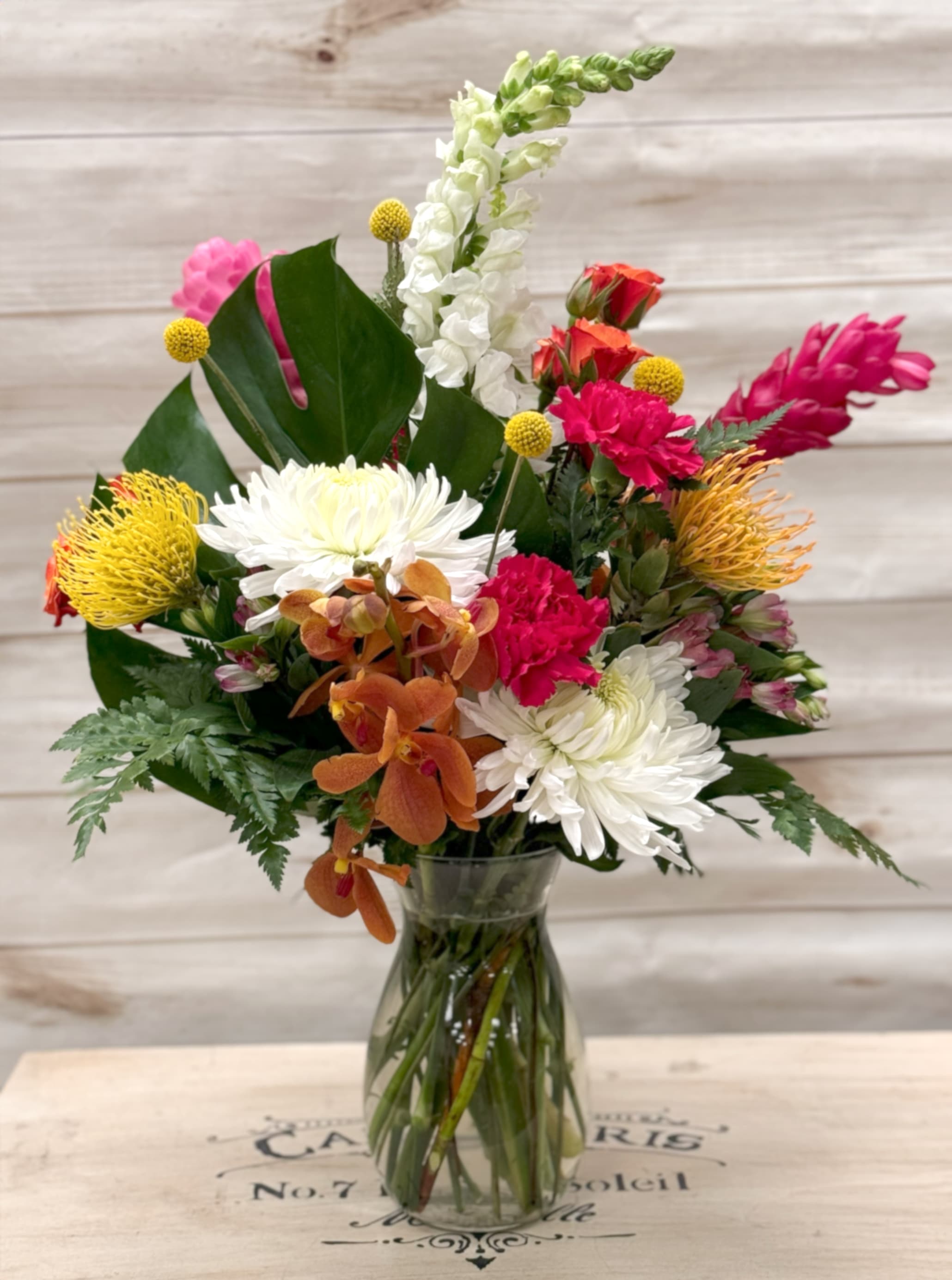 Mixed bouquet of white mums, pink carnations, yellow protea, and orange orchids in a clear glass vase