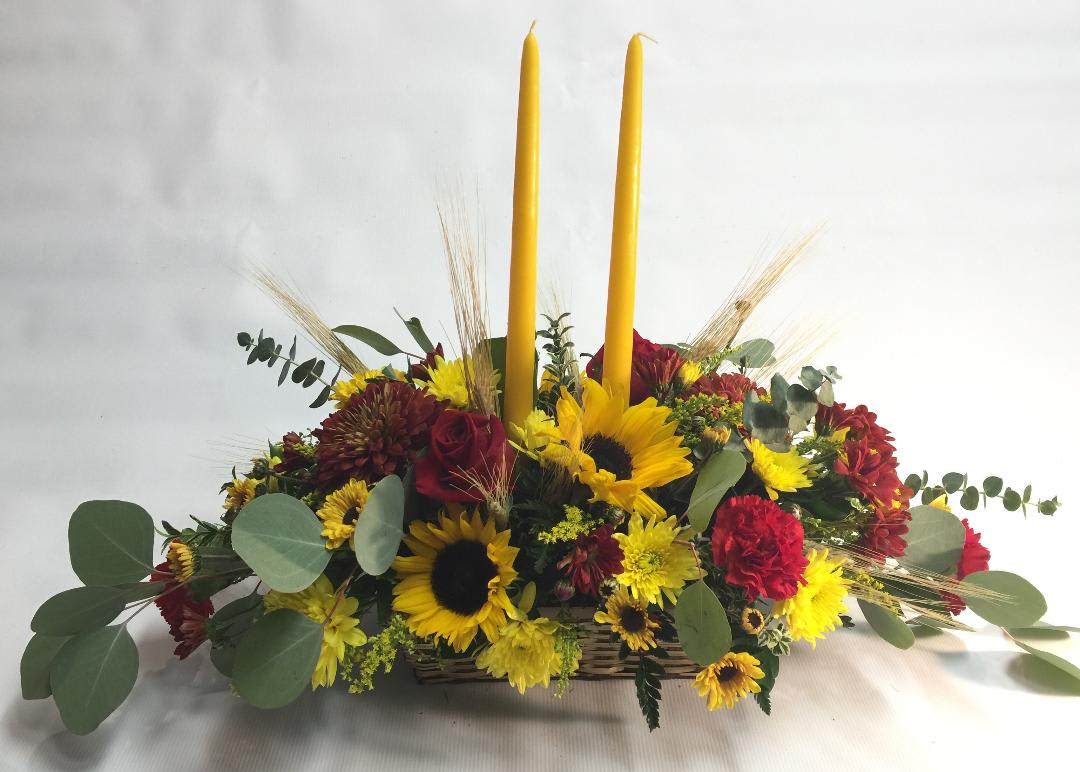 Sunflower and red flower arrangement with two tall yellow candles in a basket