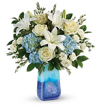White lilies and roses arranged with blue hydrangeas in a blue glass vase.