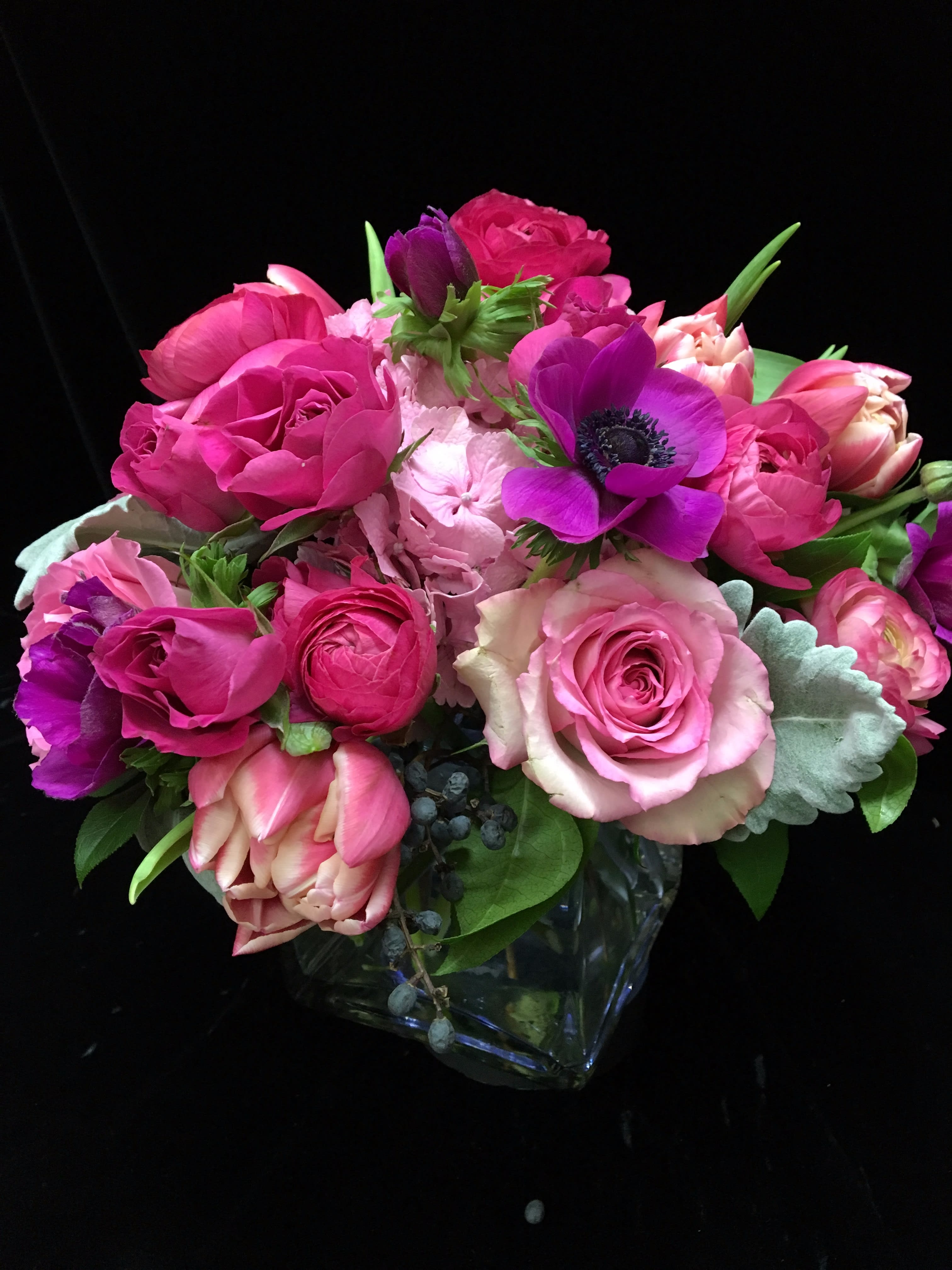 Compact arrangement of pink roses, tulips, and purple anemones in a clear glass cube vase.