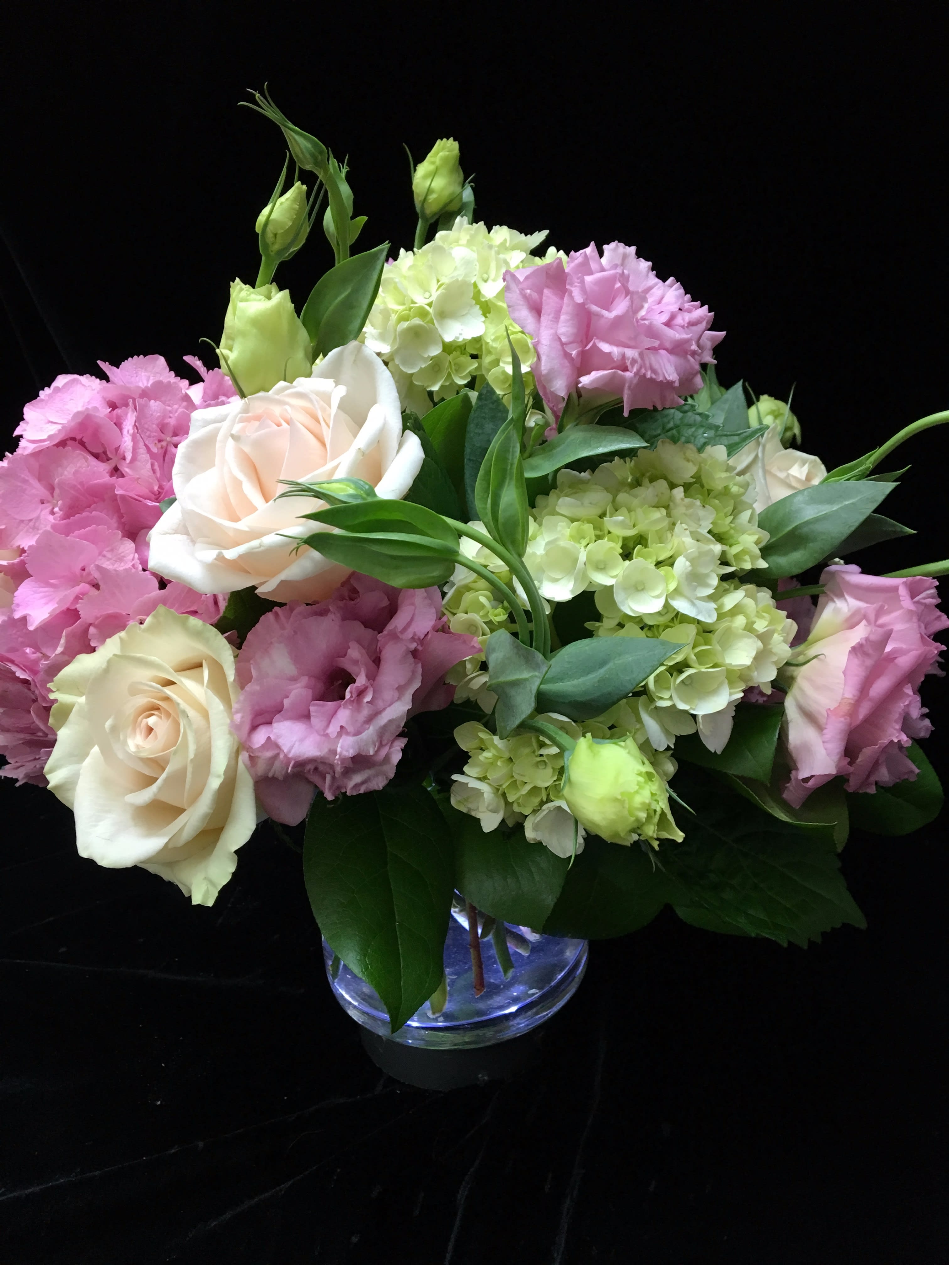 Low arrangement of cream roses, pink lisianthus, and green hydrangeas in a clear glass vase