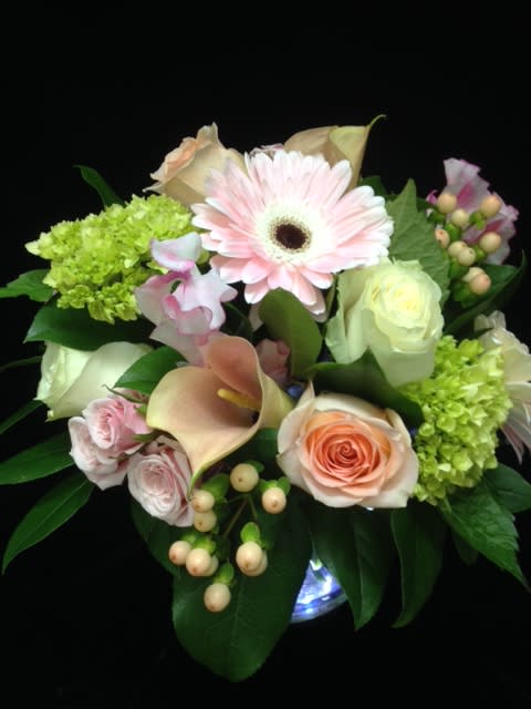Round arrangement of pale pink gerbera, roses, calla lilies, and green hydrangea in a glass vase