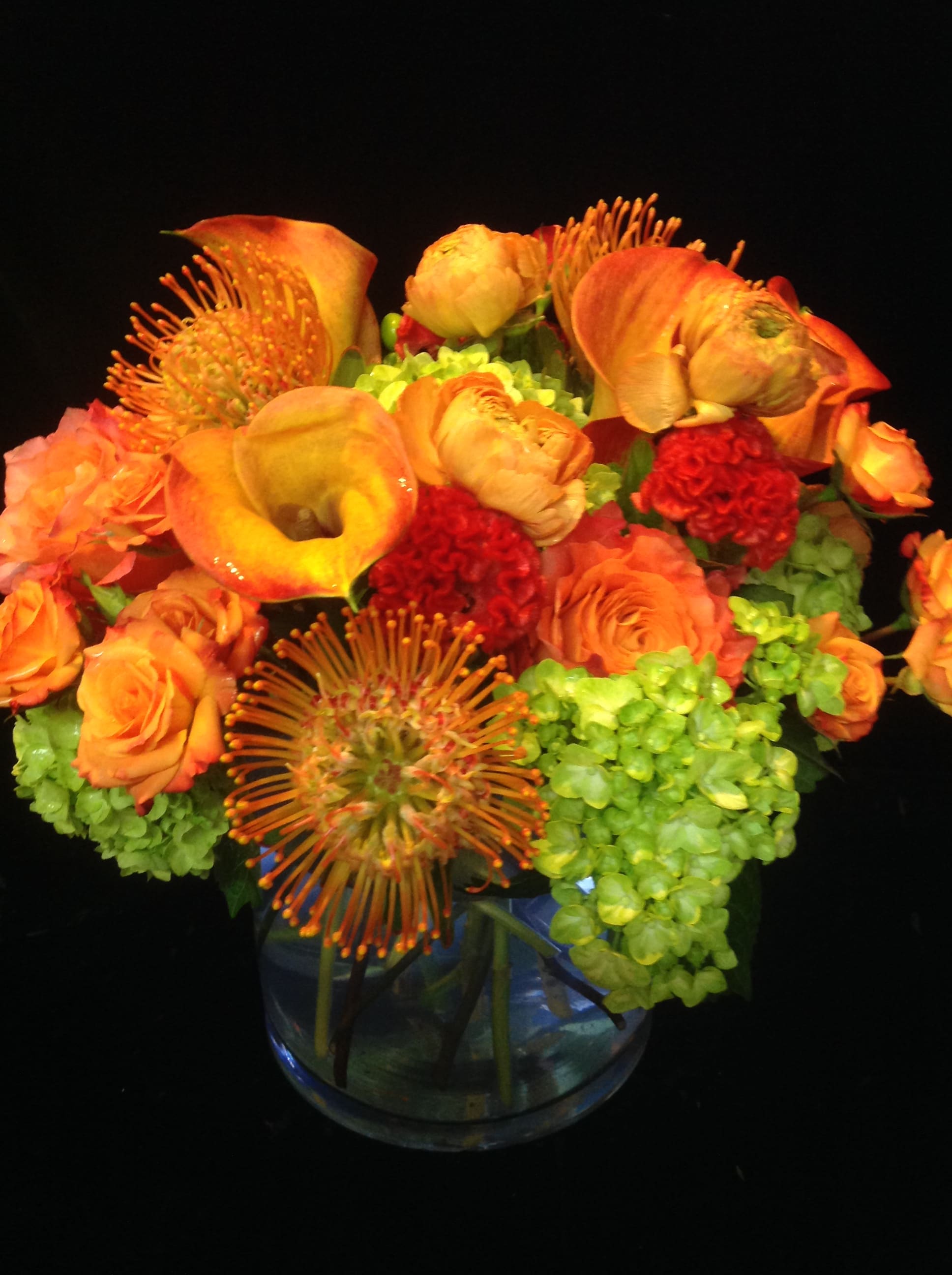Low round arrangement of orange calla lilies, roses, red blooms and green hydrangeas in a clear glass vase