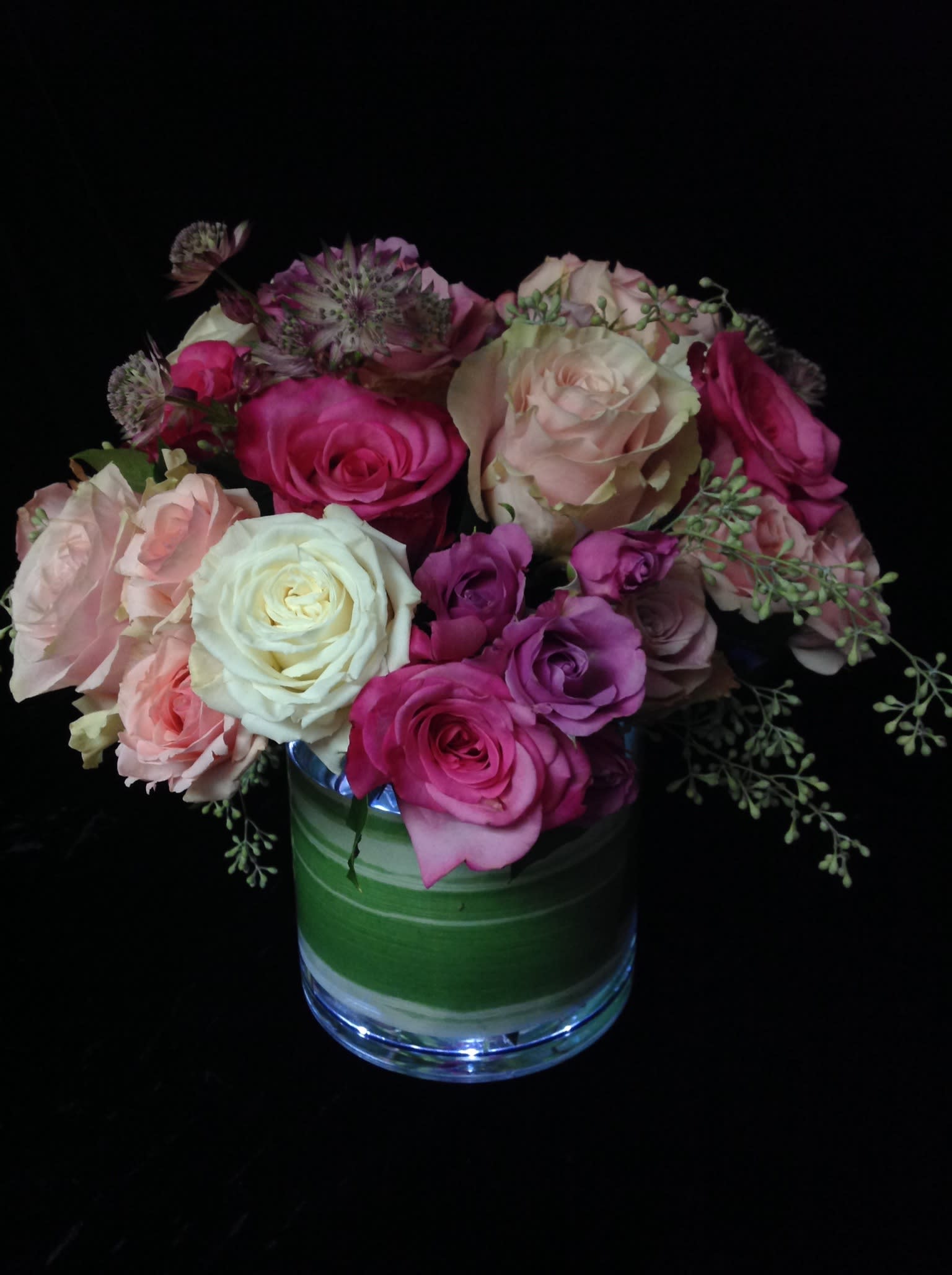 Compact arrangement of pink, peach, and cream roses in a clear glass vase with a green leaf wrap.