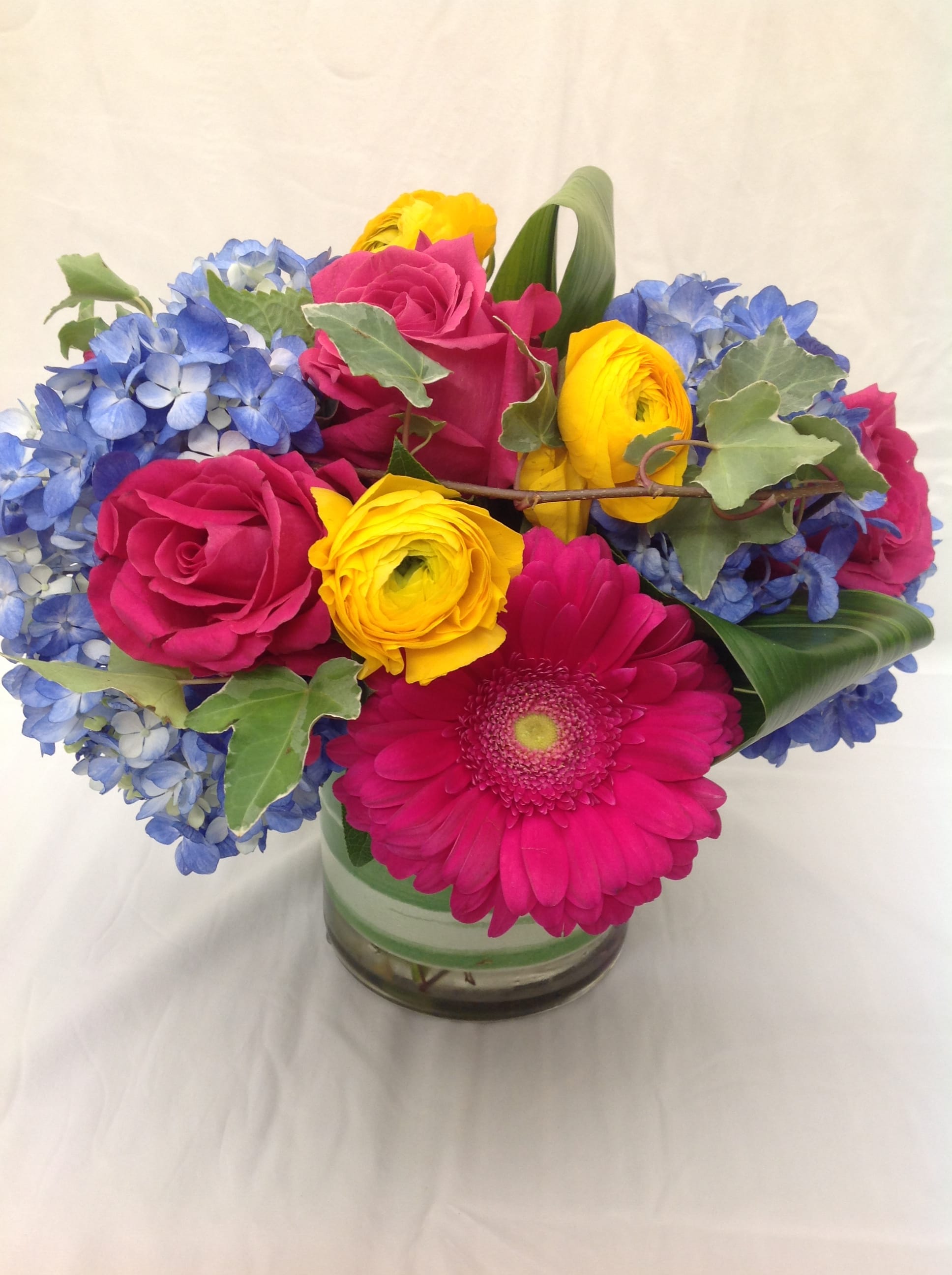 Bright arrangement of pink roses and gerbera daisy with yellow blooms and blue hydrangeas in a glass vase