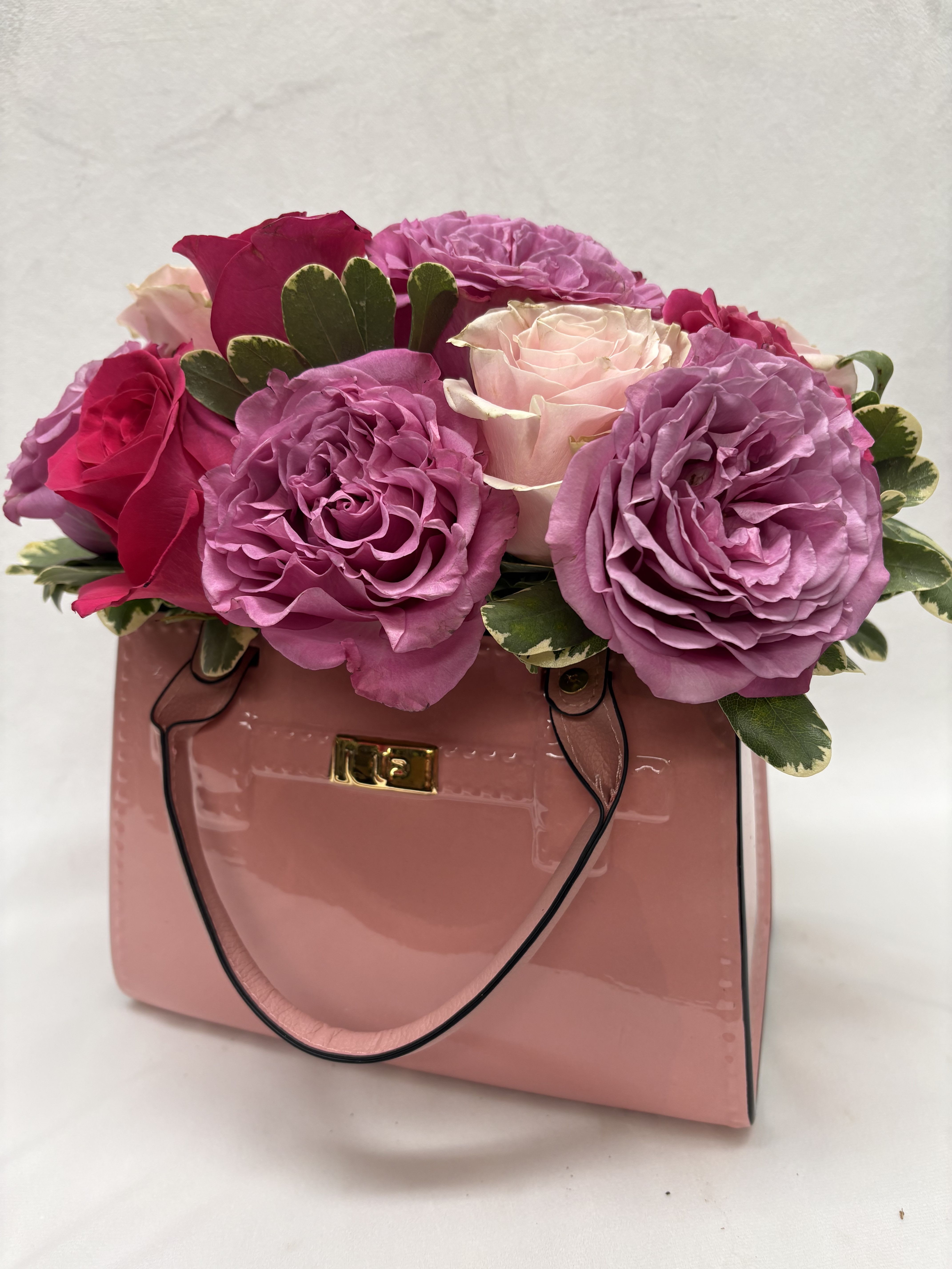 Haute Roses - A compact cluster of pink, purple, and white roses set directly into a glossy pink gift bag creates an instantly celebratory bouquet for birthdays, anniversaries, Mother's Day or simply to say "thinking of you." The rounded, tightly grouped silhouette reads feminine and romantic-perfect for love and romance, a new baby visit, graduation, or a springtime table centerpiece that makes the recipient feel truly seen. The arrangement features hot-pink, blush and lavender roses arranged across the top of a soft-pink gift bag with black cord handles and a gold-tone logo plaque. Variegated foliage peeks between the blooms to add contrast and texture, giving the design a fresh, polished look that photographs beautifully and travels well.  A great choice for holiday gifting, quinceañera parties, thank-you gestures or just because moments, this design arrives ready to display-no vase required. For best enjoyment, keep away from direct heat and refresh the floral foam or water source as directed to maintain freshness through the week.