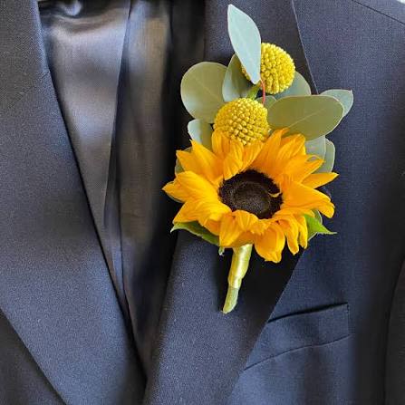 Sunflower boutonnière  - A single bright sunflower takes center stage in this sunflower boutonnière, a sunny accent perfect for wedding parties, prom, or any dressy event needing a cheerful pop of color. Pinned to a dark suit lapel, the compact boutonnière reads formal yet joyful-an ideal choice when the occasion calls for warmth and celebration and you want the person you're honoring to feel effortlessly radiant.  The design pairs the bold yellow sunflower with two round yellow accent blooms and pale green foliage, finished with subtle dark (black) elements that anchor the look against navy or charcoal suits. Small and tailored, it sits close to the lapel for a neat silhouette; the mixed textures-from the sunflower's textured center to the soft greenery-create visual contrast that's visible even at a distance.  Perfect for groomsmen, prom dates, or anyone taking part in a sunny-themed event, this boutonnière sends a message of cheer and confidence. Order to match tux rental timelines; keep it cool and dry before the event and pin it on just before photos to preserve freshness.