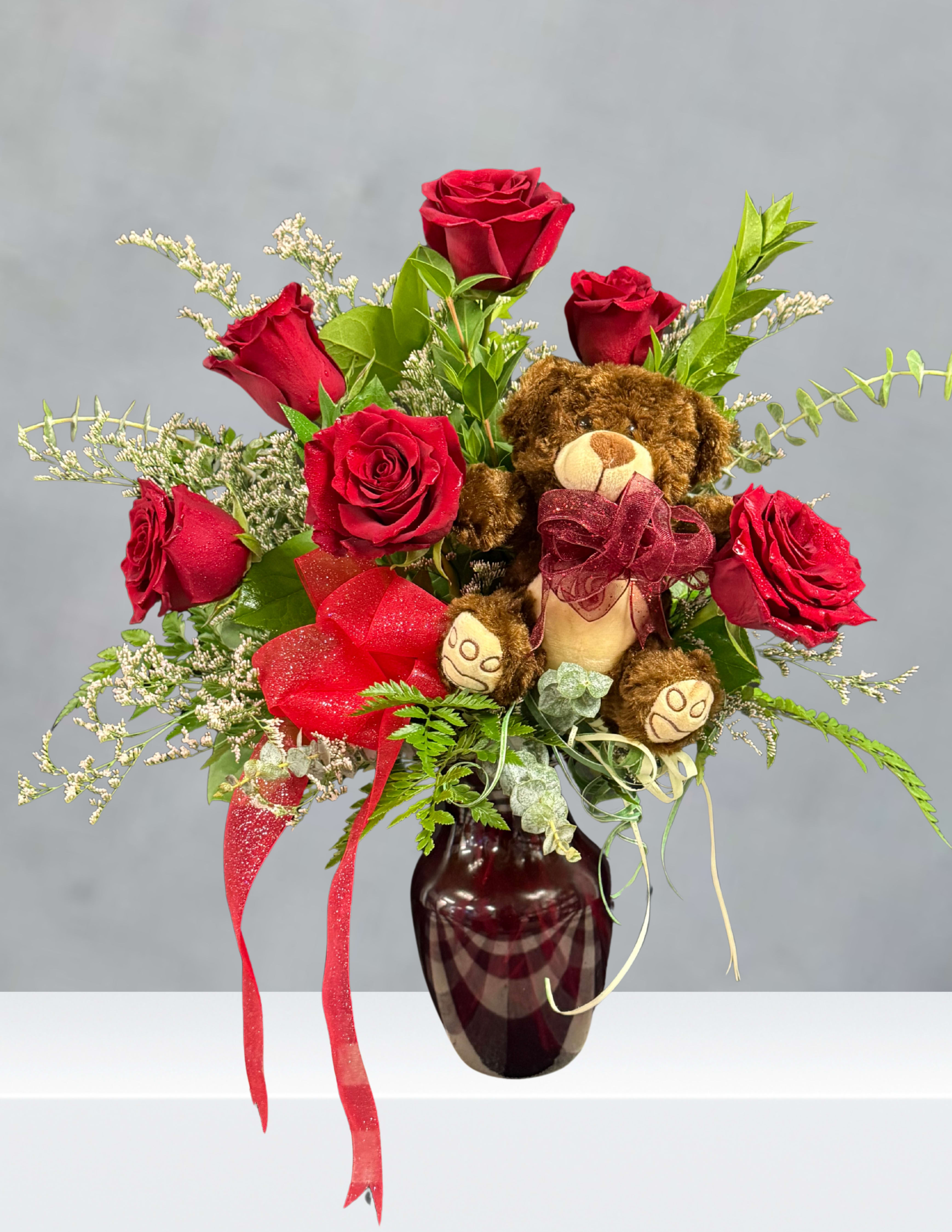 Red roses arranged in a vase with a teddy bear and ribbon accents