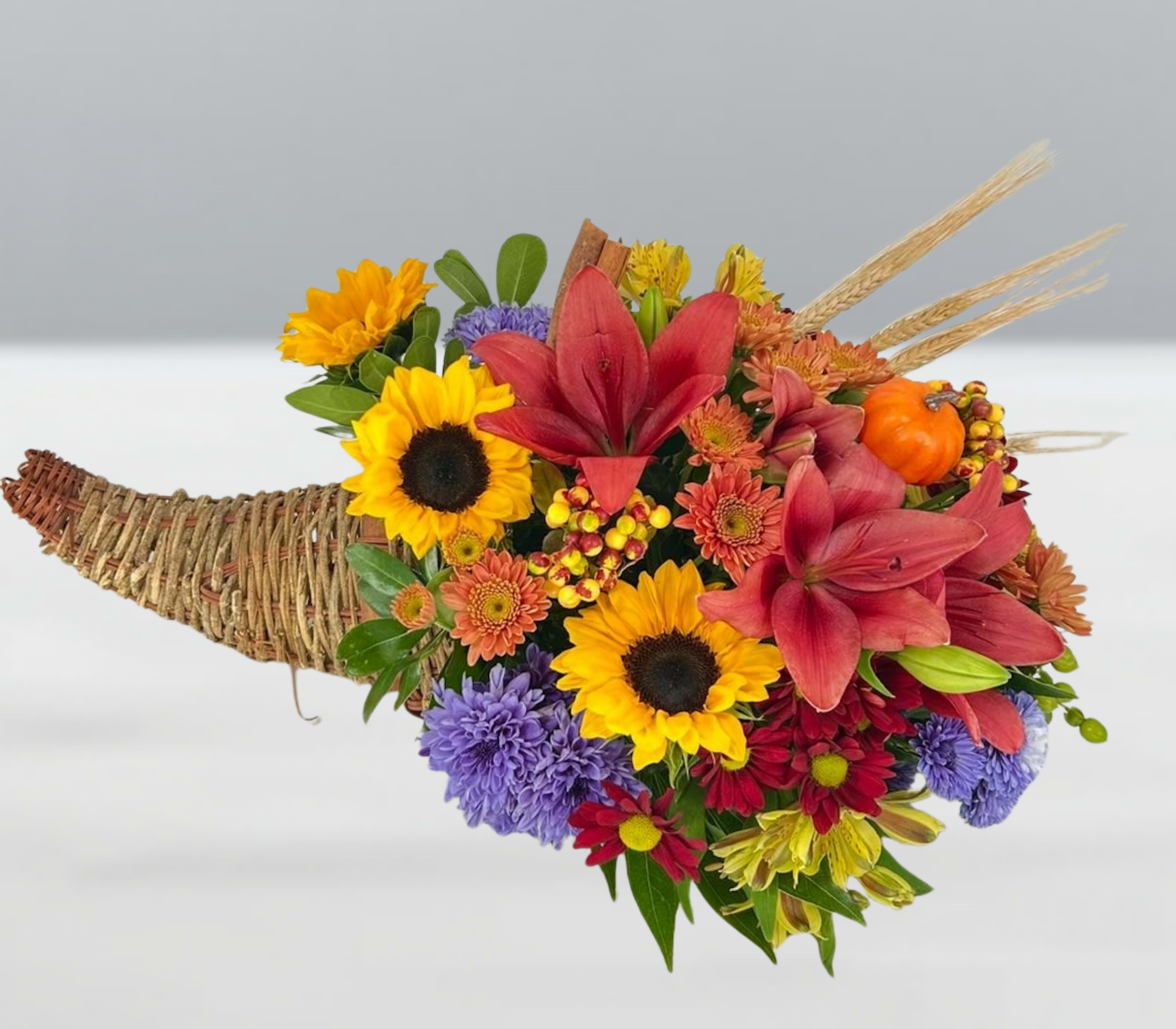 Autumn rhapsody -  Autumn Rhapsody  A symphony of fall’s finest—this cornucopia bursts with red lilies, golden sunflowers, orange mums, and violet asters, harmonized with wheat stalks, berries, and a playful pumpkin accent. Each bloom sings of abundance, warmth, and celebration, creating a visual melody that captures the heart of harvest season. Autumn Rhapsody is more than a centerpiece—it’s a crescendo of color and gratitude, ready to grace your table or gift with joy.