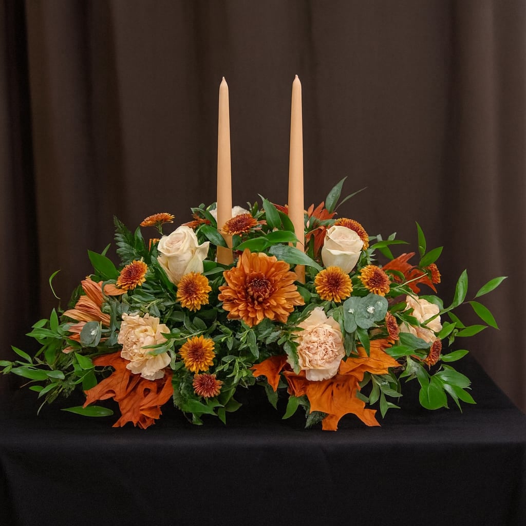 The giving flame - ️ The Giving Flame  A centerpiece of gratitude and glow—this lush autumn arrangement features rust-hued chrysanthemums, creamy roses, peach carnations, and golden daisies nestled among eucalyptus and Italian ruscus. Two taper candles rise from the heart of the design, casting a soft, flickering light that evokes togetherness and tradition. The Giving Flame is perfect for Thanksgiving tables, heartfelt gatherings, or any moment meant to feel warm, generous, and beautifully lit.