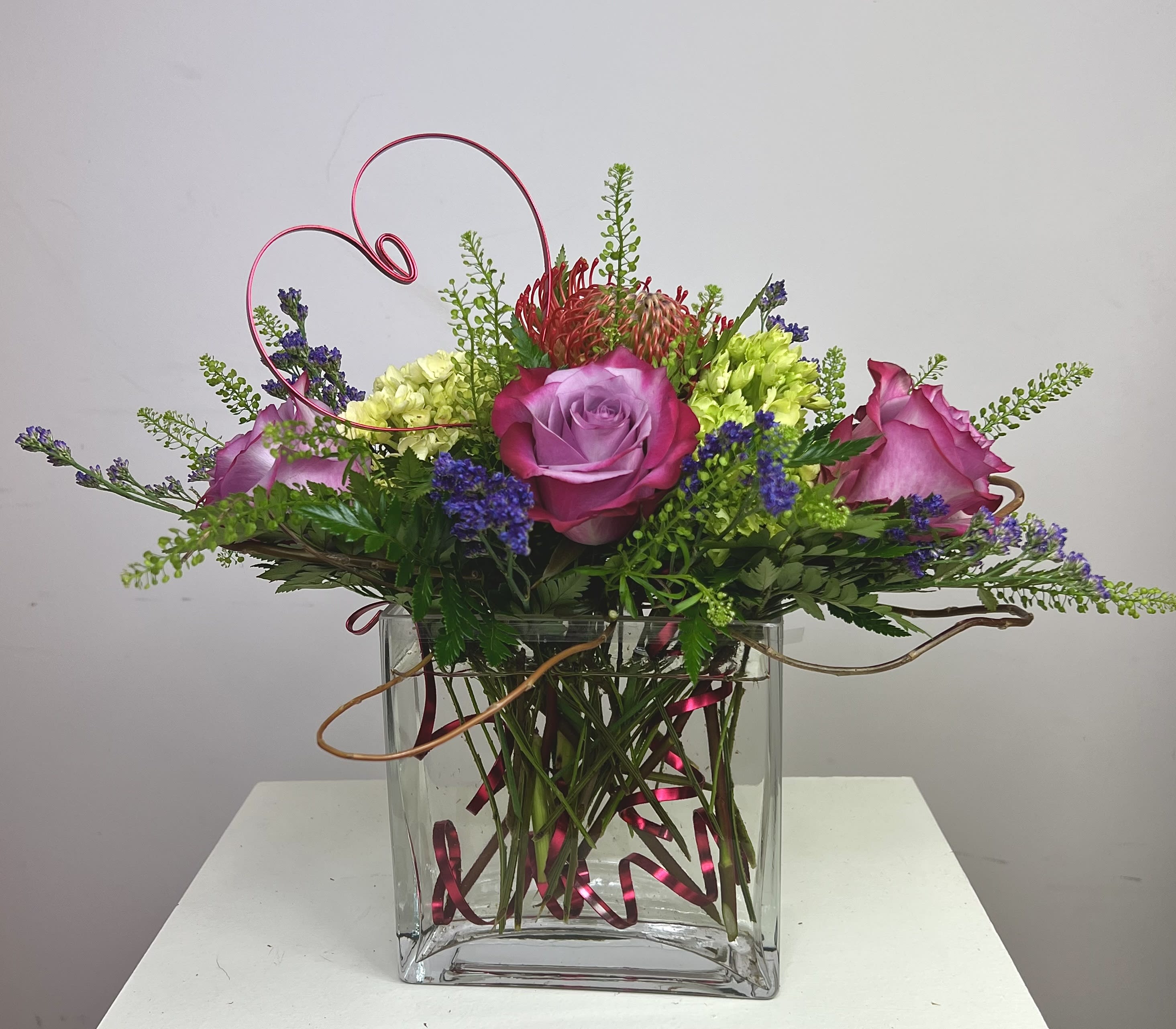 Pink and purple roses arranged in a clear square vase with curly wire accents