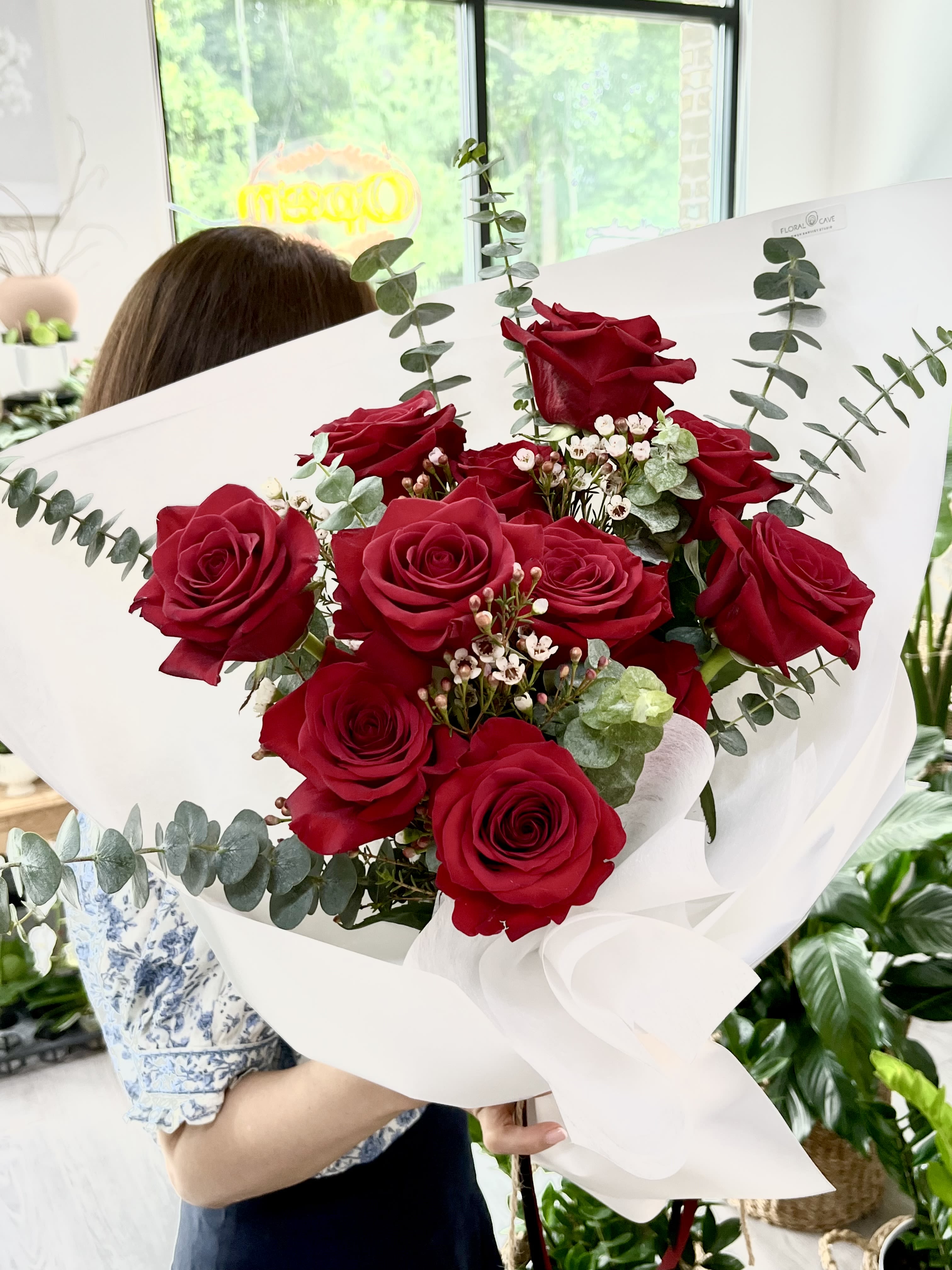 Luxury Dozen Red Rose Elegance Bouquet - A dense cluster of deep red roses arranged in a rounded, hand-tied bouquet creates an instant feeling of romance-perfect as an anniversary roses bouquet, a Valentine's Day gift, or a love-and-romance gesture that makes the person you're thinking of feel truly cherished. The rich red blooms are set against airy eucalyptus and delicate white filler blossoms, giving the arrangement a modern yet classic silhouette that reads elegant from every angle.  Wrapped in crisp white paper and finished with a soft, wide ribbon, the presentation is clean and polished-ready to hand over without fuss. The cool gray-green of eucalyptus lifts the intensity of the red petals while the small white flowers soften the composition, creating texture and a balanced color story in red, white, and green.  This is the kind of bouquet that marks meaningful moments or brightens an ordinary day: a romantic surprise, a birthday for a partner, or a thoughtful "I'm thinking of you." Hand-tied for a comfortable, natural hold; care is simple-place in fresh water, recut stems, and enjoy the roses as they open over several days.