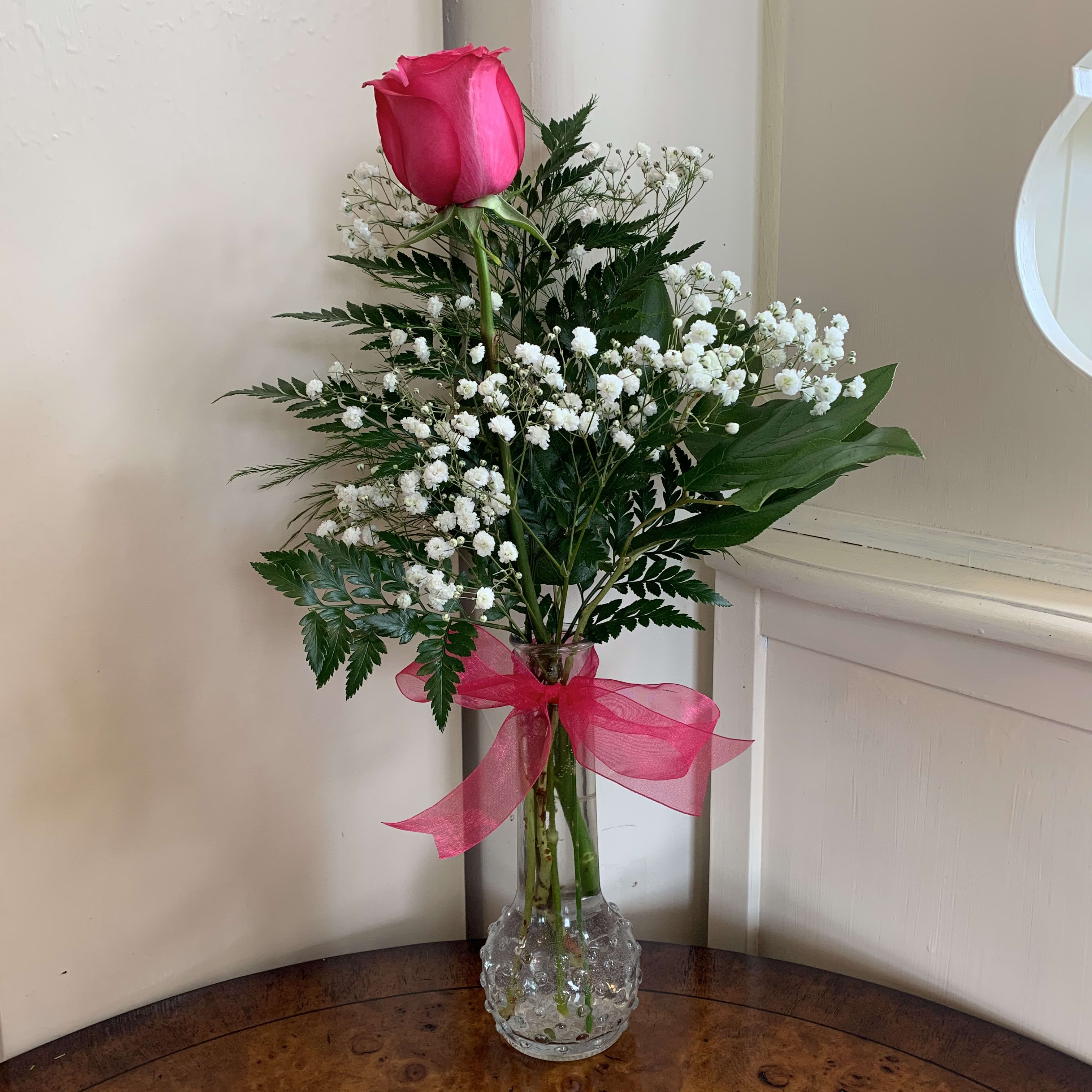 Single pink rose with baby's breath in a clear glass vase tied with a pink ribbon.