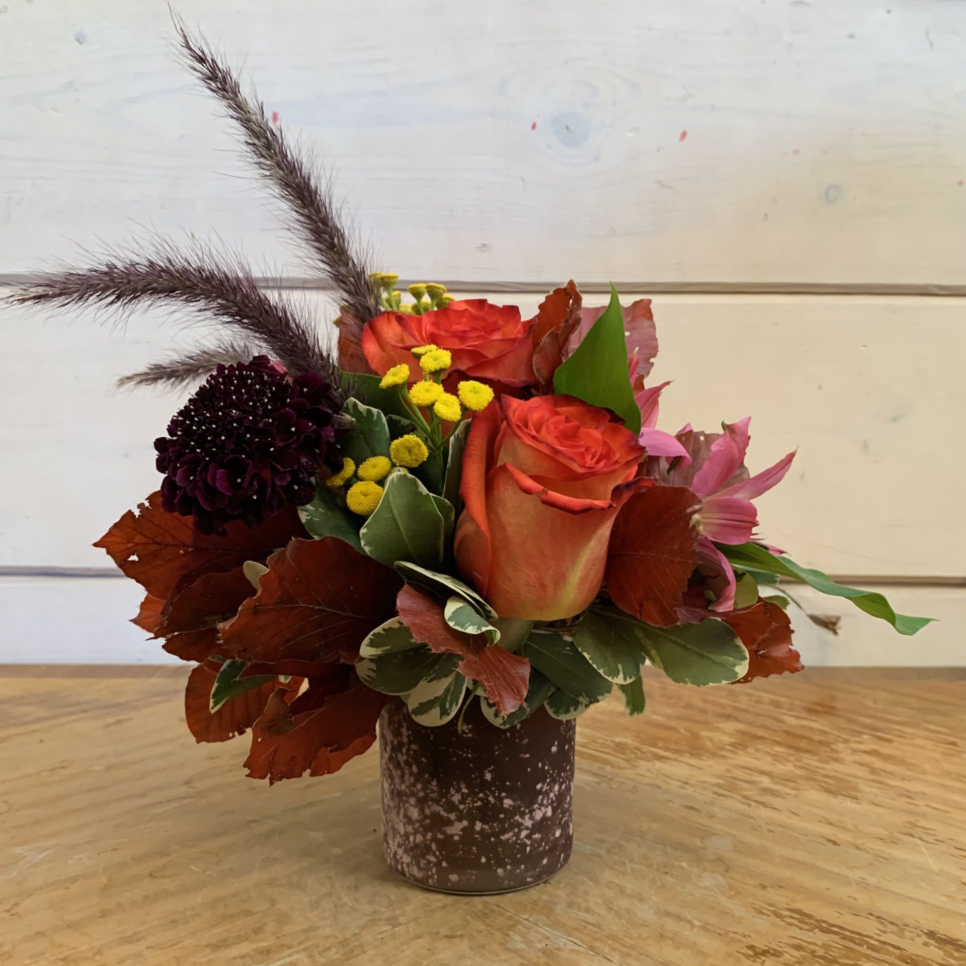 Orange roses in a small brown vase with dark accent flowers