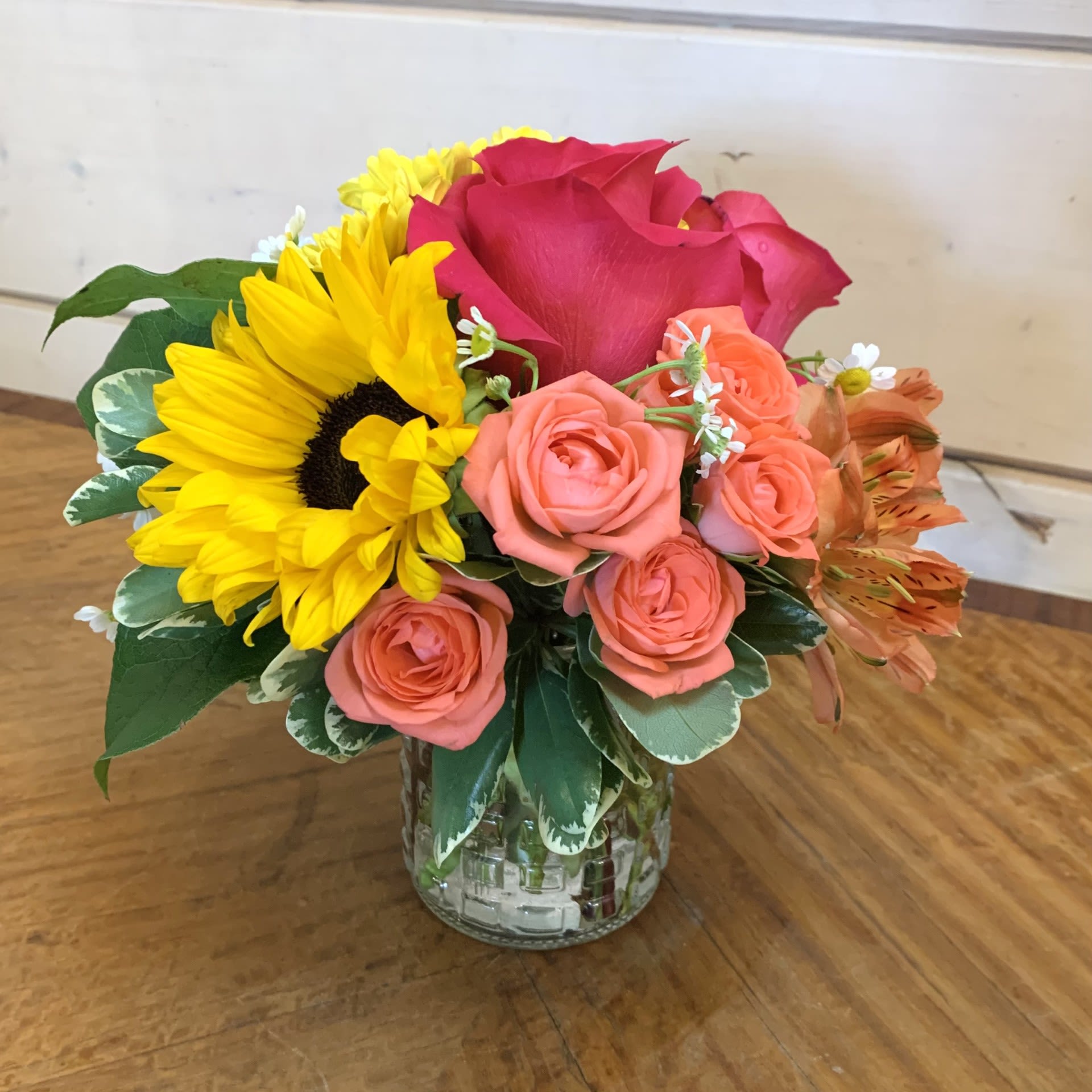 A Little Summer - A petite arrangement of hot pink roses and coral spray roses with a sunflower and viking daisies in a petite glass vase with orange alstroemeria and chamomile. Approximately 6" by 6".