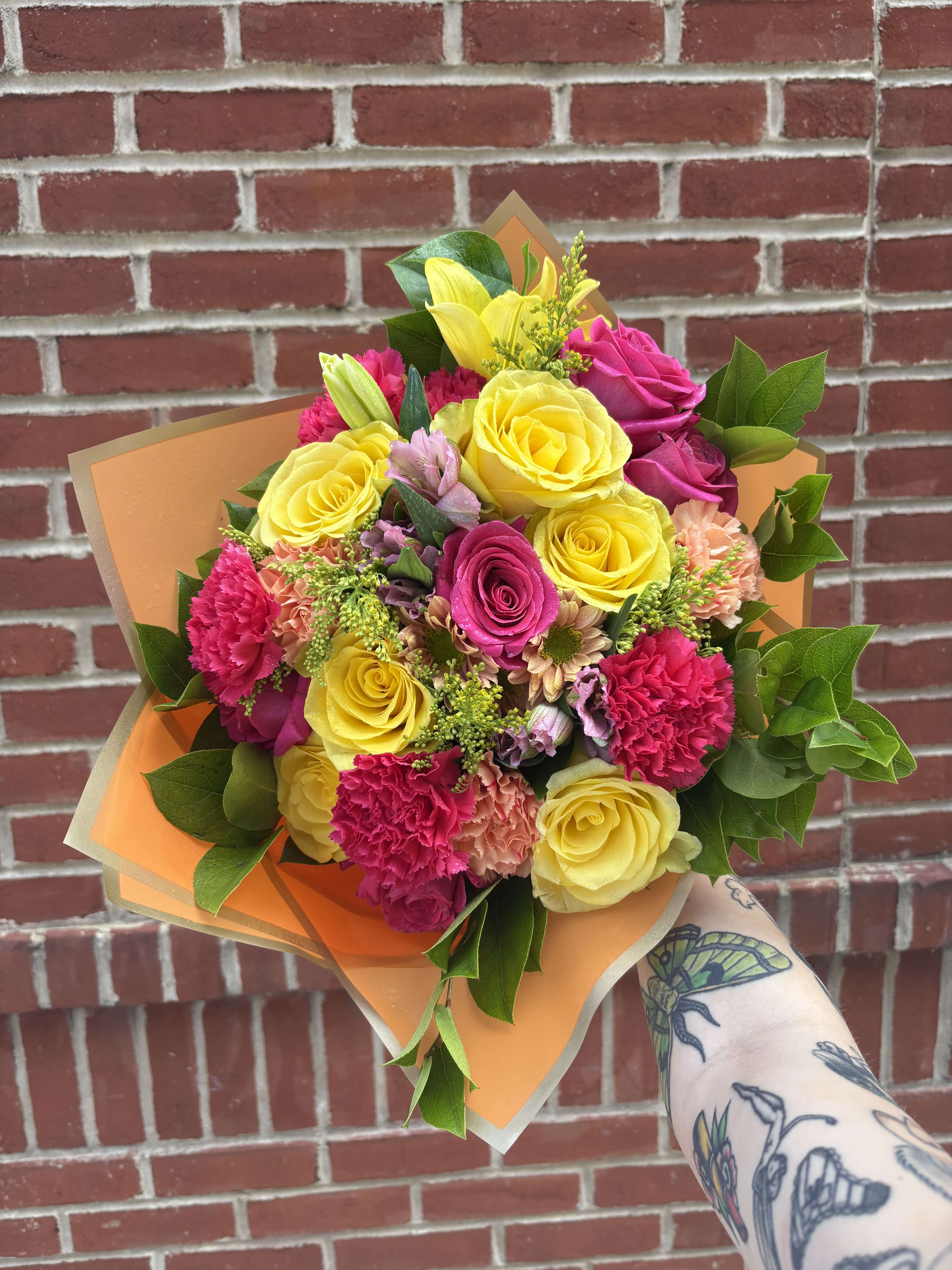 Handheld bouquet of yellow and pink roses with carnations in orange wrap