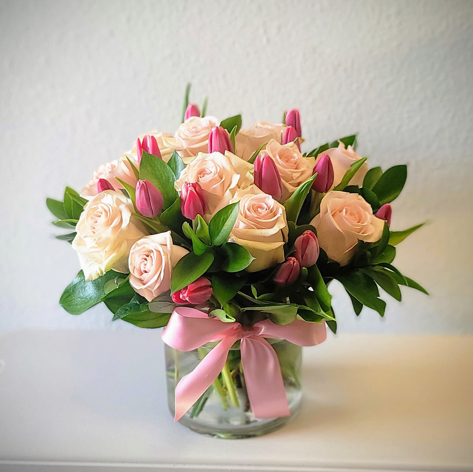Blush roses and pink tulips arranged in a clear glass vase with a pink ribbon.
